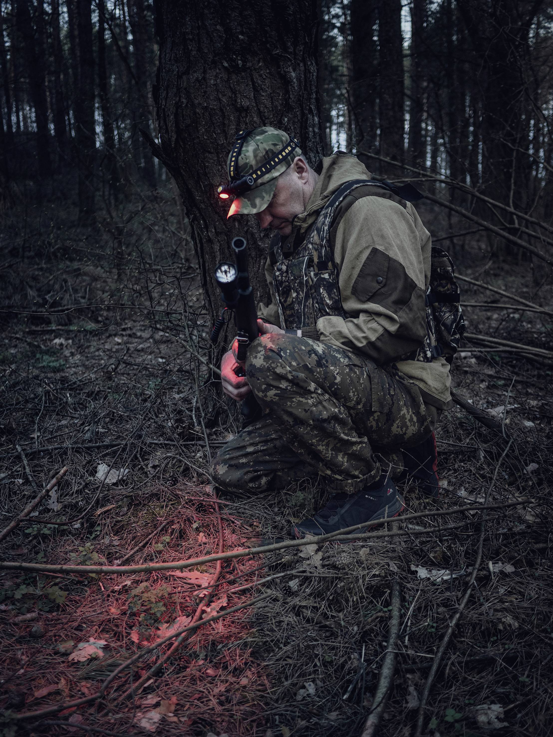 Osoba v maskáčoch kľačí v lese so červeným svetlom. Pripravená na opierke pre zbraň, sústredeným pohľadom upretým na zem. Lovecká alebo nočná aktivita.