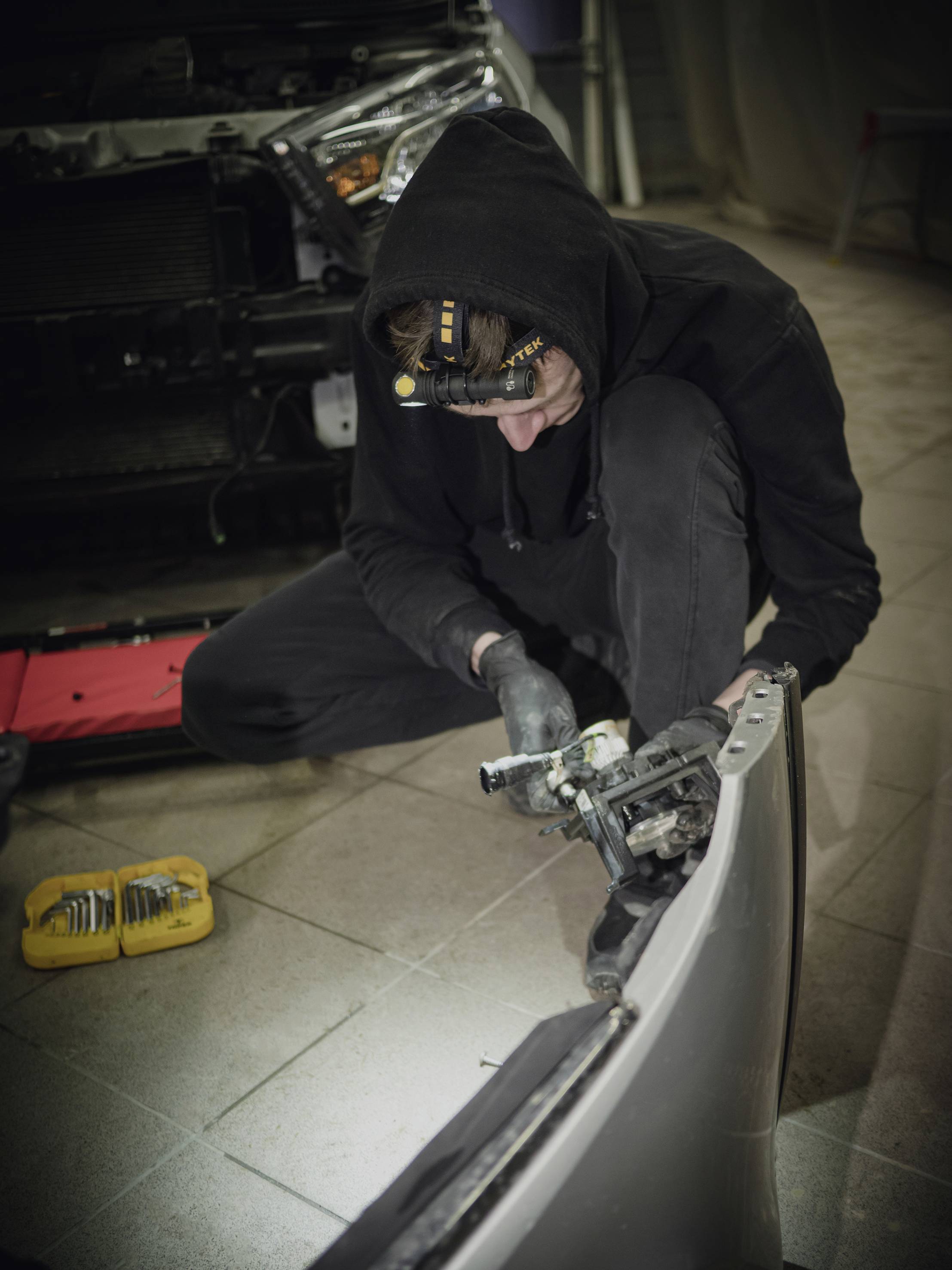Osoba v čiernom pracovnom oblečení opravuje automobilovú súčiastku v dielni. V pozadí je čiastočne vidieť automobil.