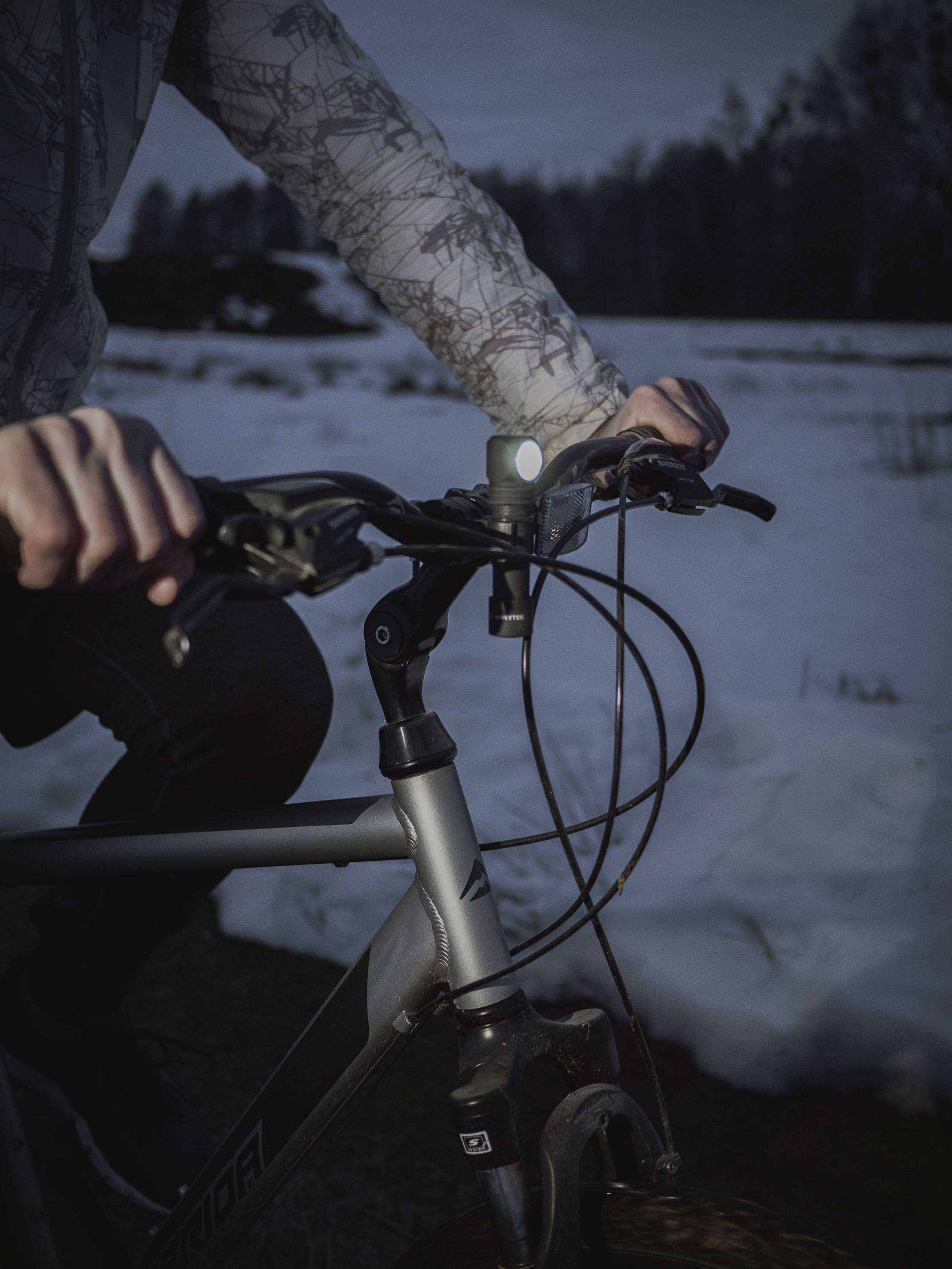Osoba jazdí na bicykli počas súmraku na zasneženej ceste. Zaostrené na riadidlá a ruky, pozadie rozmazané so snehom.