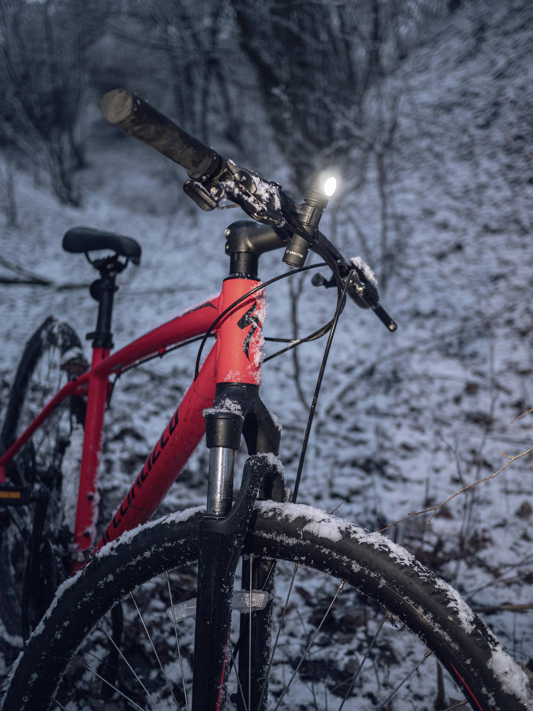 Červený bicykel stojí na zasneženej lesnej ceste. Predné svetlo svieti a osvetľuje okolie.