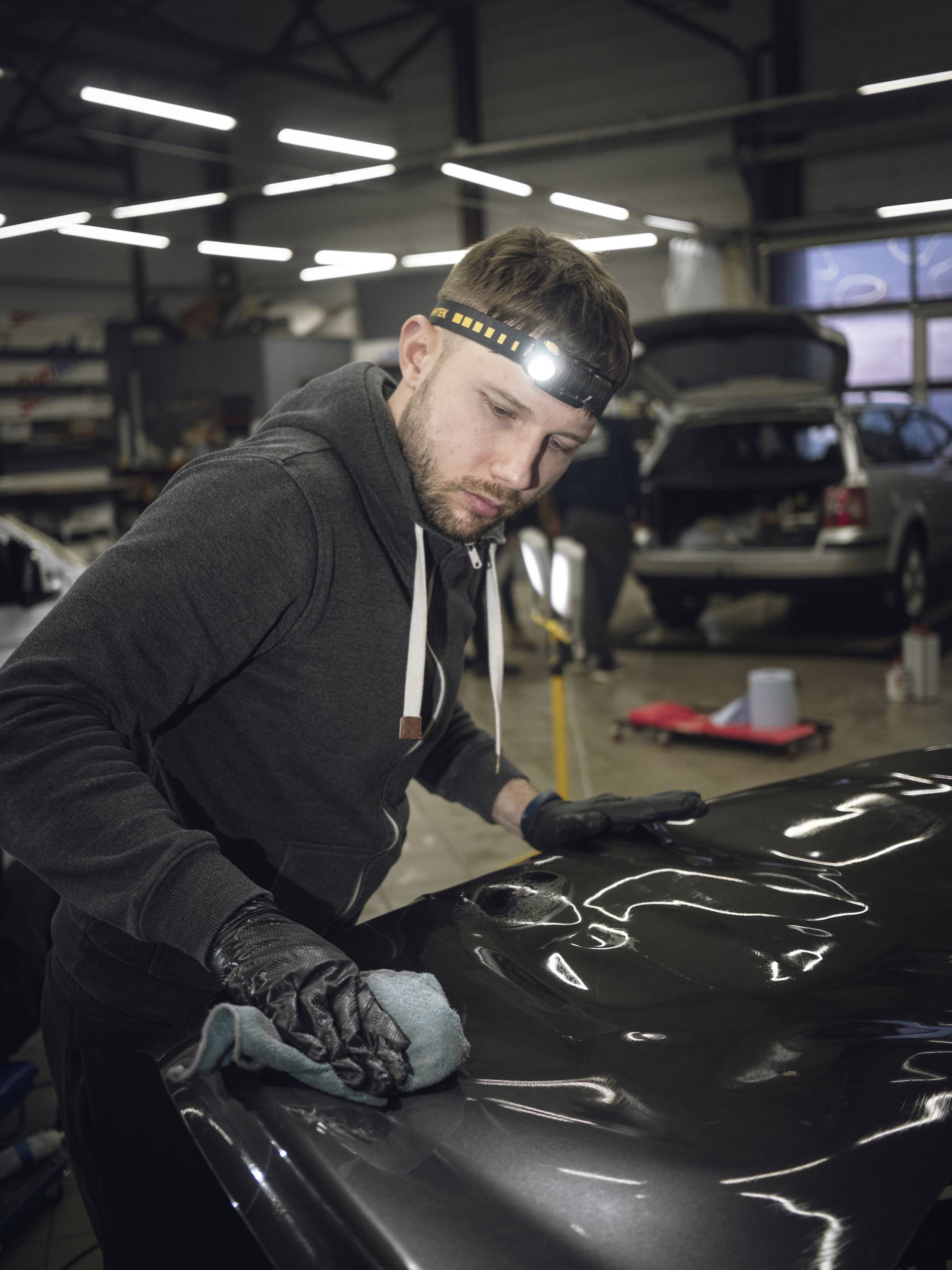 Mechanik leští kapotu automobilu v dielni, pričom má na hlave čelovku, aby vylepšil povrchovú úpravu laku.