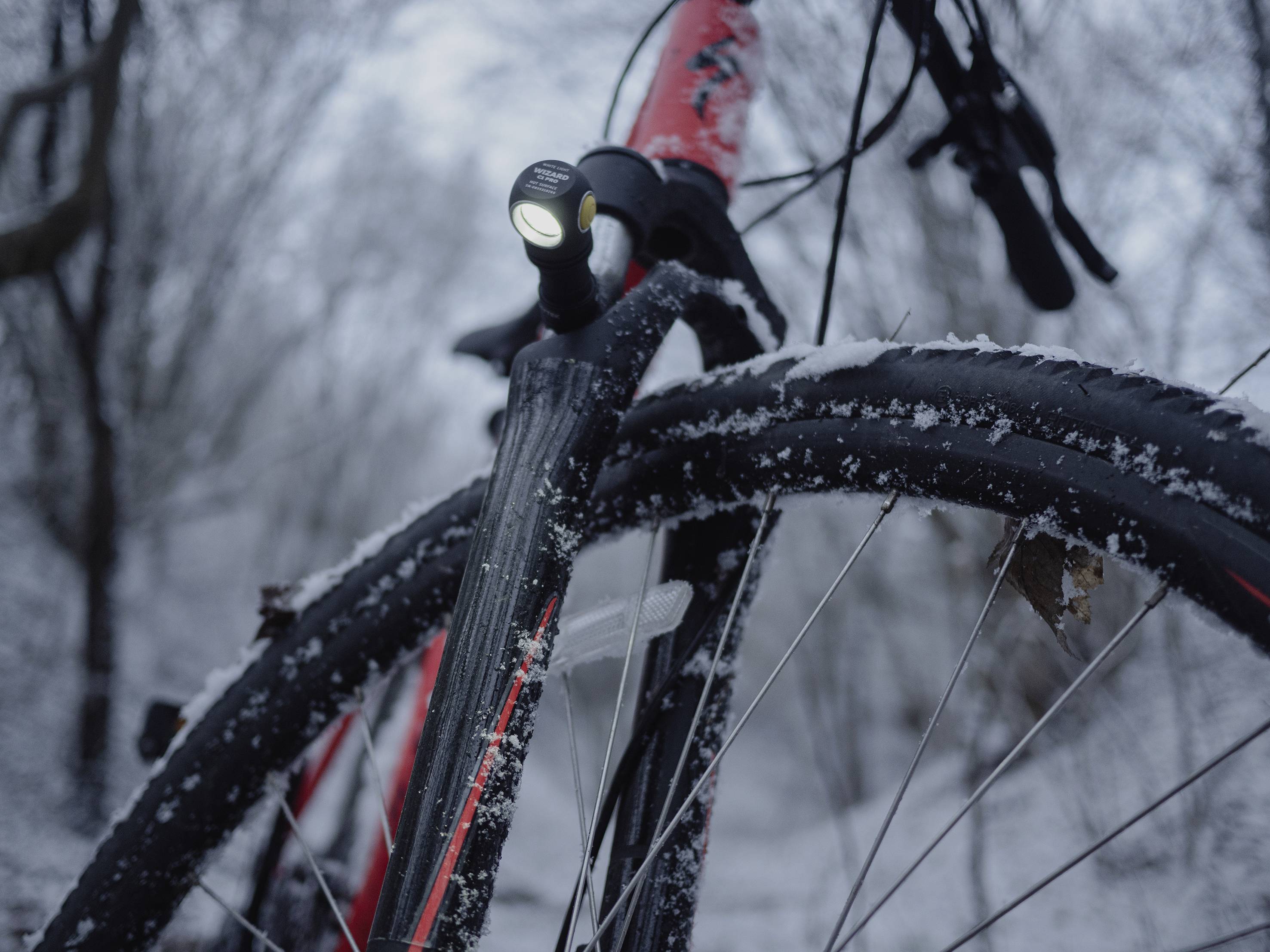 Bicykel v snehu so zapnutým predným svetlom, obklopený zimnou krajinou. Hlavný fokus na prednom kolese a svetle.