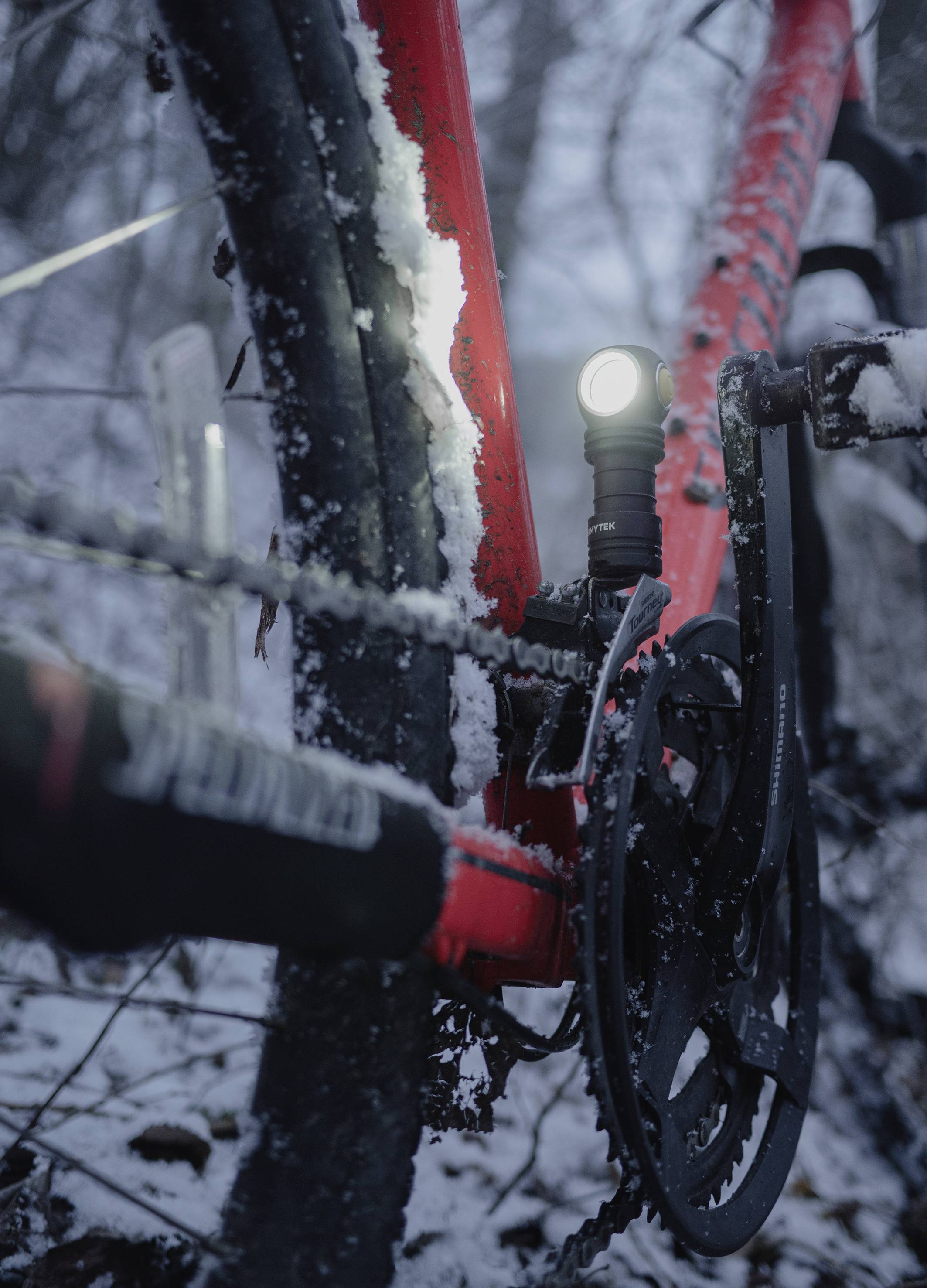 Detailný záber červeného bicykla v snehu so zapnutým svetlom. Reťaz a ozubené koleso sú viditeľné. Zimné prostredie.