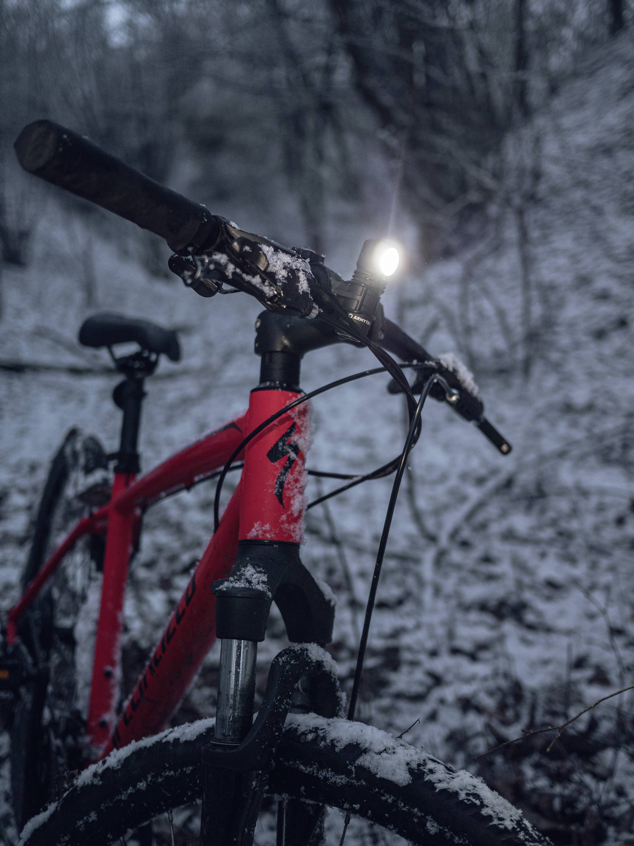 Červený bicykel v zasneženom lese, smerujúci dopredu. Predné koleso a rám sú pokryté snehom, svetlo svieti jasne.