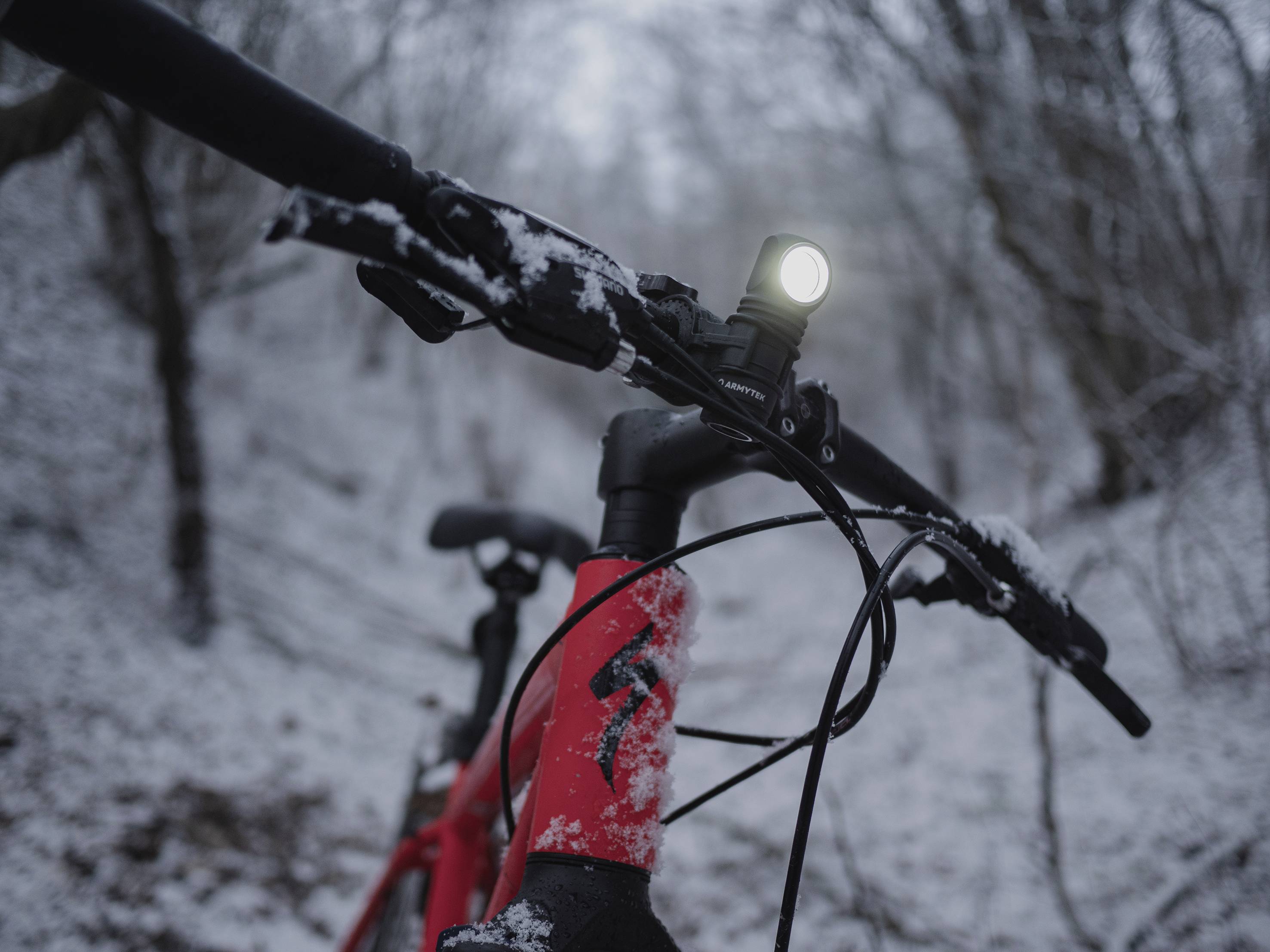 Červený bicykel so zapnutým predným svetlom stojí v zasneženom lese. Riadidlá sú pokryté snehom.