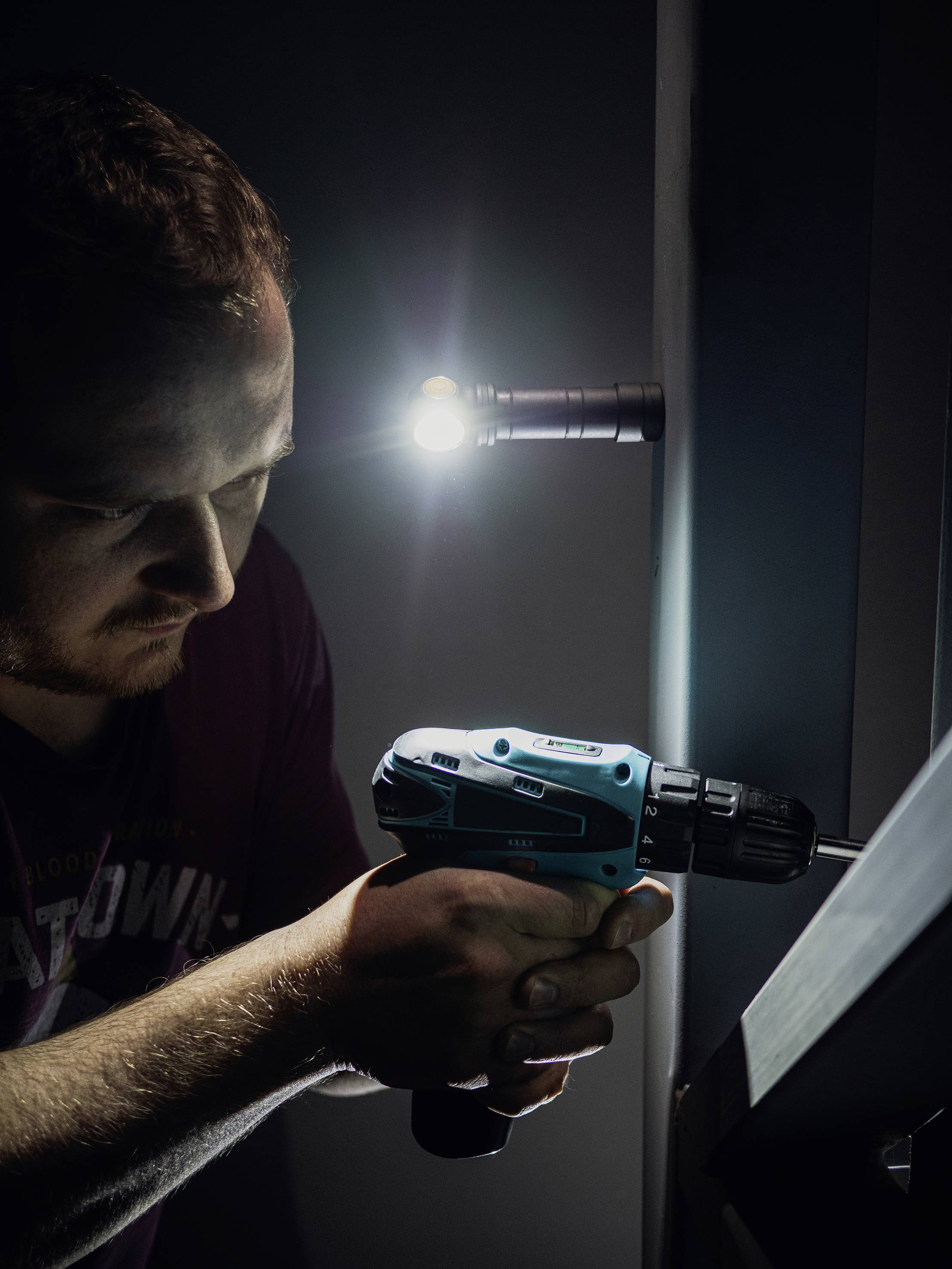 Muž používa elektrickú vŕtačku, aby zaskrutkoval skrutku do steny. Lampa osvetľuje pracovný priestor.