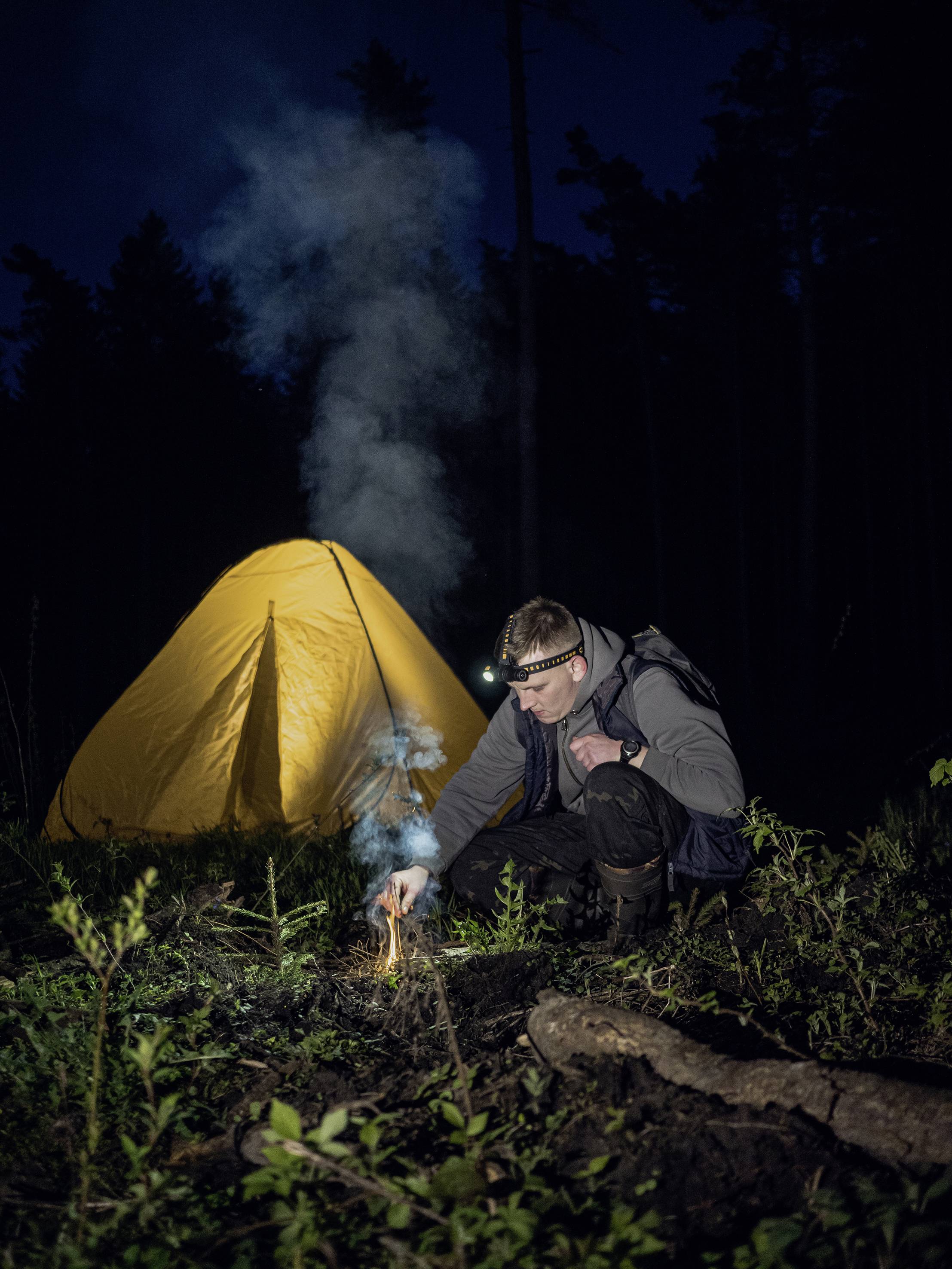 Osoba zapaľuje malý oheň pred žltým stanom v nočnom lese. Má čelovku a je oblečená v outdoorovom oblečení.