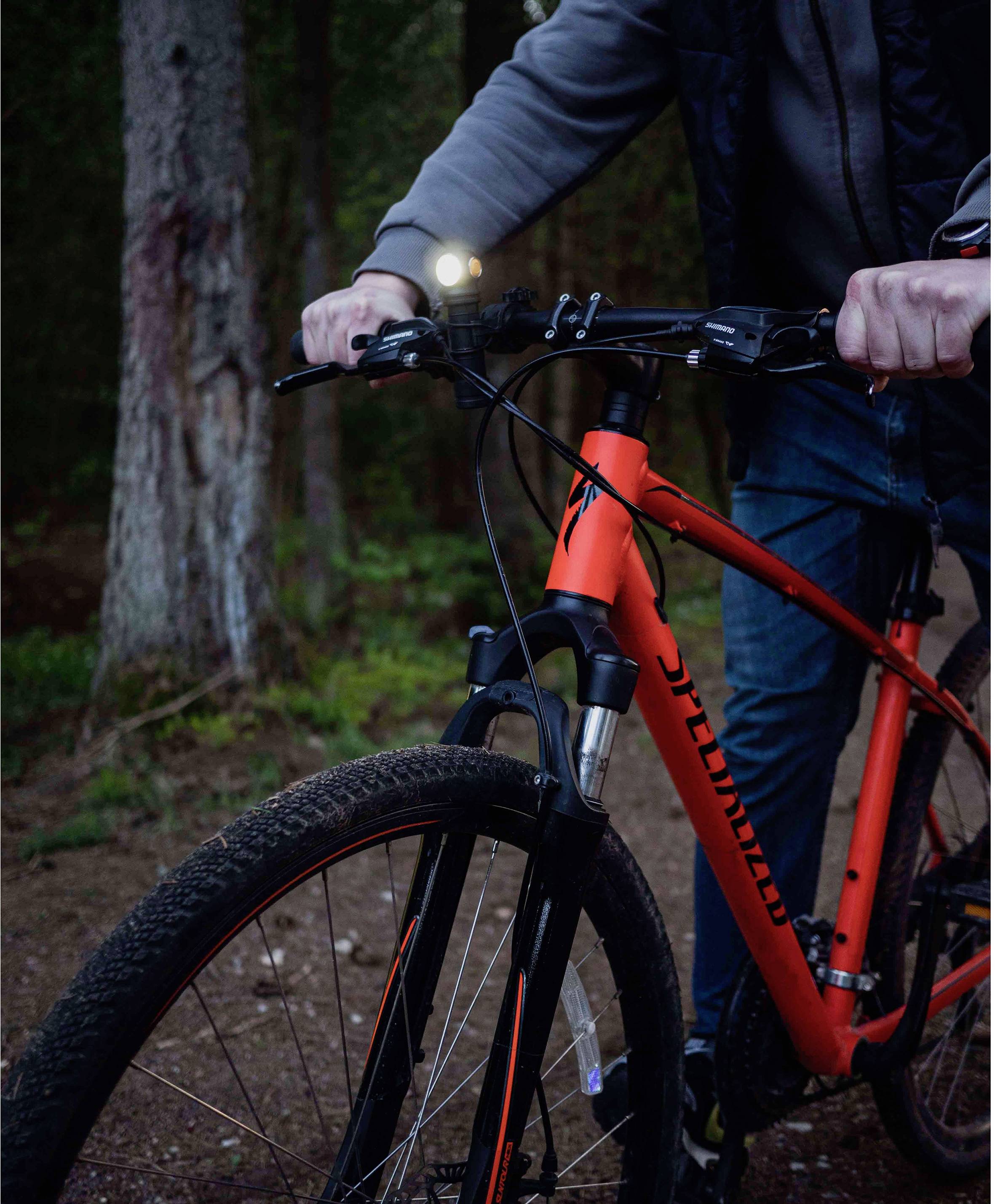 Osoba jazdí na červenom bicykli po lesnej ceste. Svetlo bicykla je zapnuté.