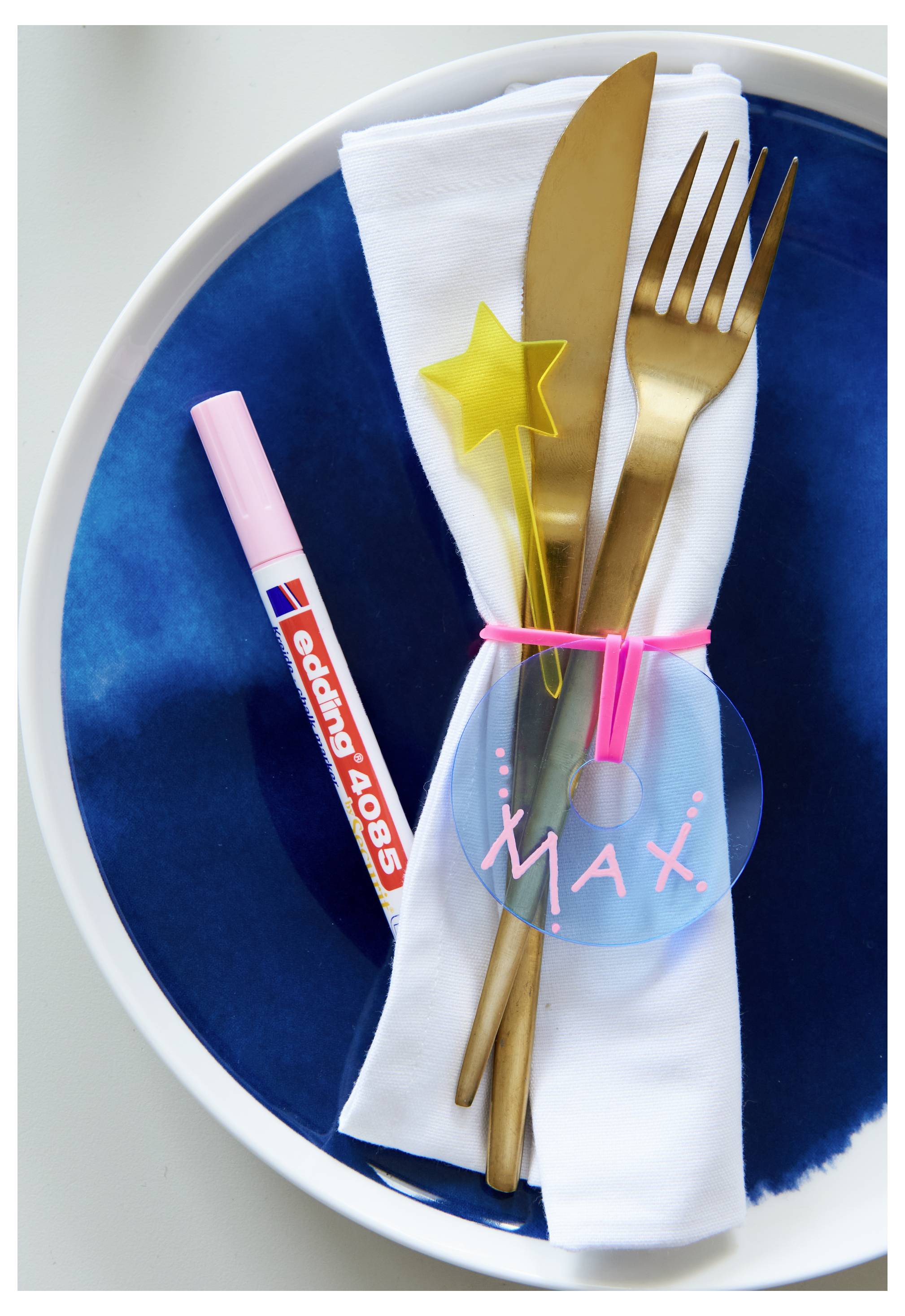 Stolovanie s zloženom bielym obrúskom na modrom tanieri, zviazanom menovkou "Max" a ružovou stužkou, plus zlaté príbory a miešadlo v tvare hviezdy; vedľa neho spočíva pero s ružovými pruhmi.