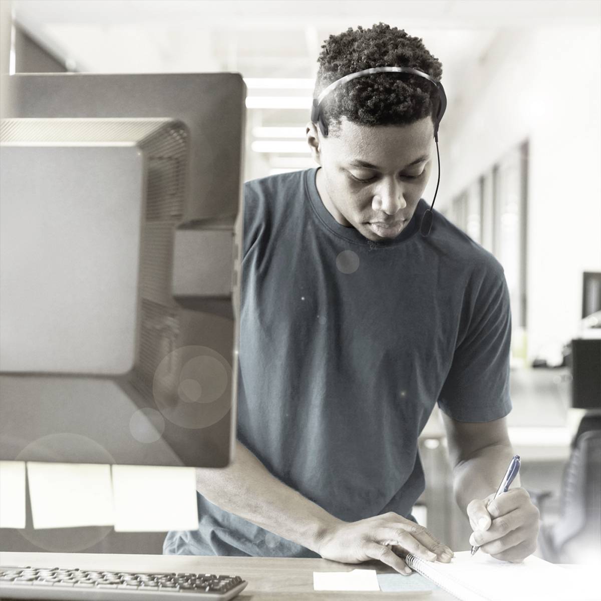 Muž s headsetom pracuje na počítači a robí si poznámky. V pozadí je kancelária.