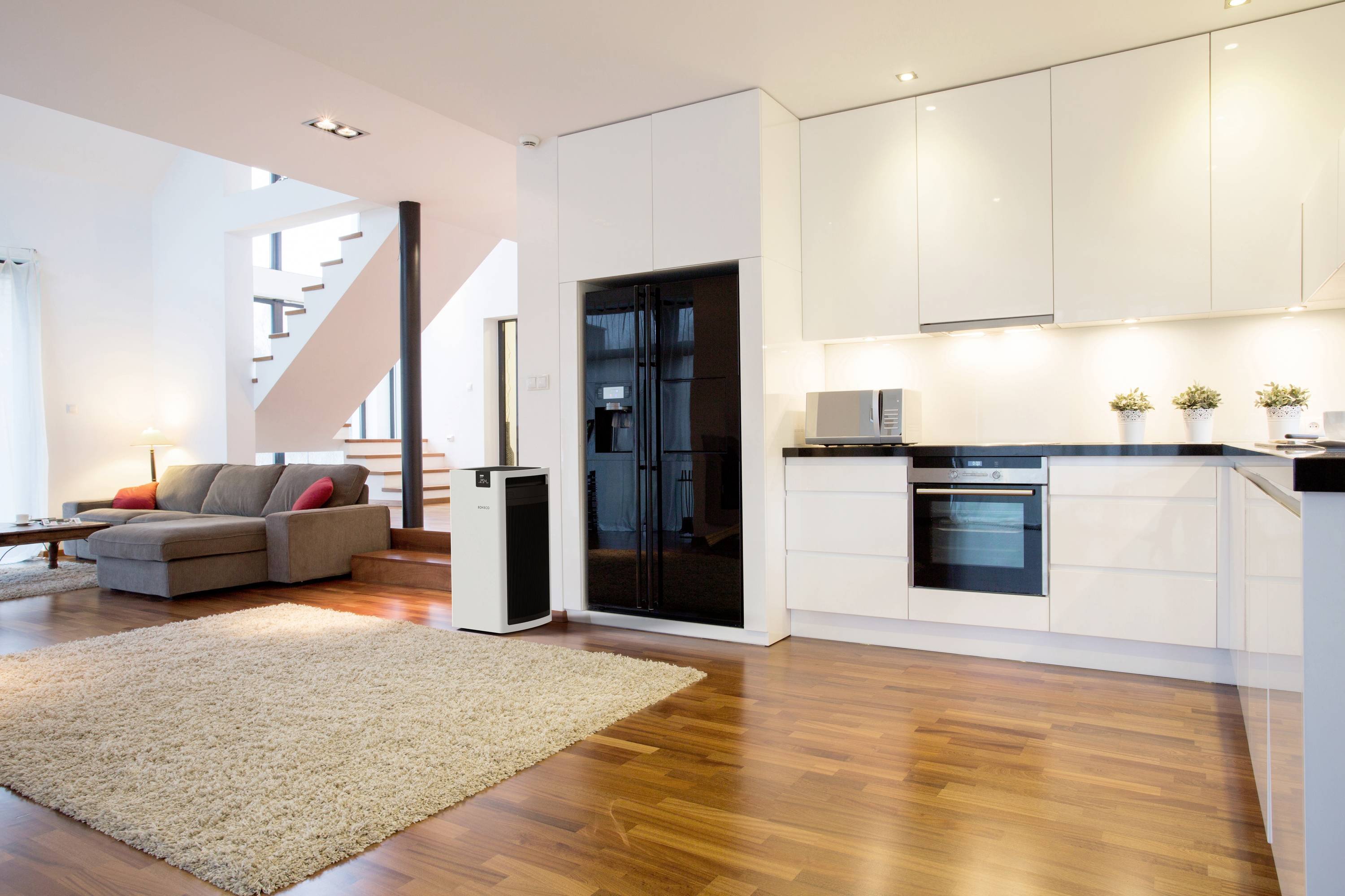 Svetlá, moderná kuchyňa s obývacím priestorom s drevenou parketovou podlahou, bielou vstavanou kuchynskou linkou, čiernou chladničkou, kobercom a útulným sedacím kútom v pozadí.