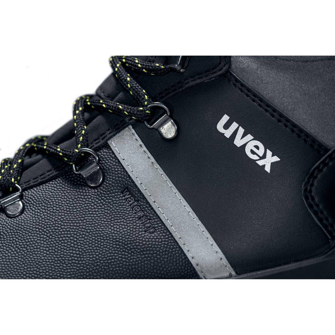 Detail blízkej fotografie čiernej bezpečnostnej topánky so značkovým logom „uvex". Topánka má čierno-žlté šnúrky a šedú aplikáciu.