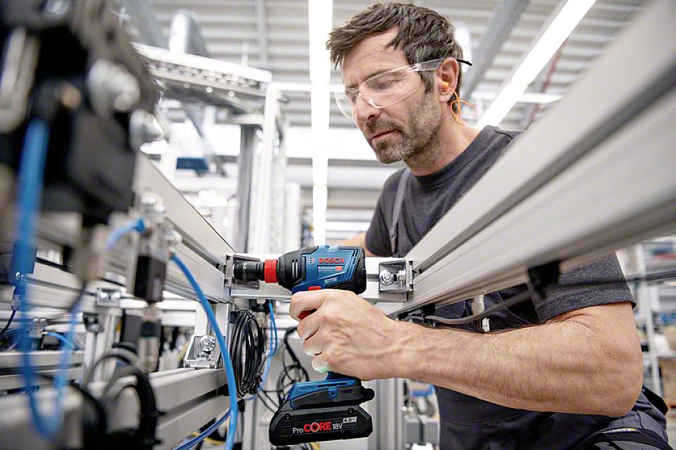 Muž v ochranných okuliaroch pracuje s akumulátorovým skrutkovačom na priemyselnom montážnom projekte, obklopený kovovými rámami a káblami.