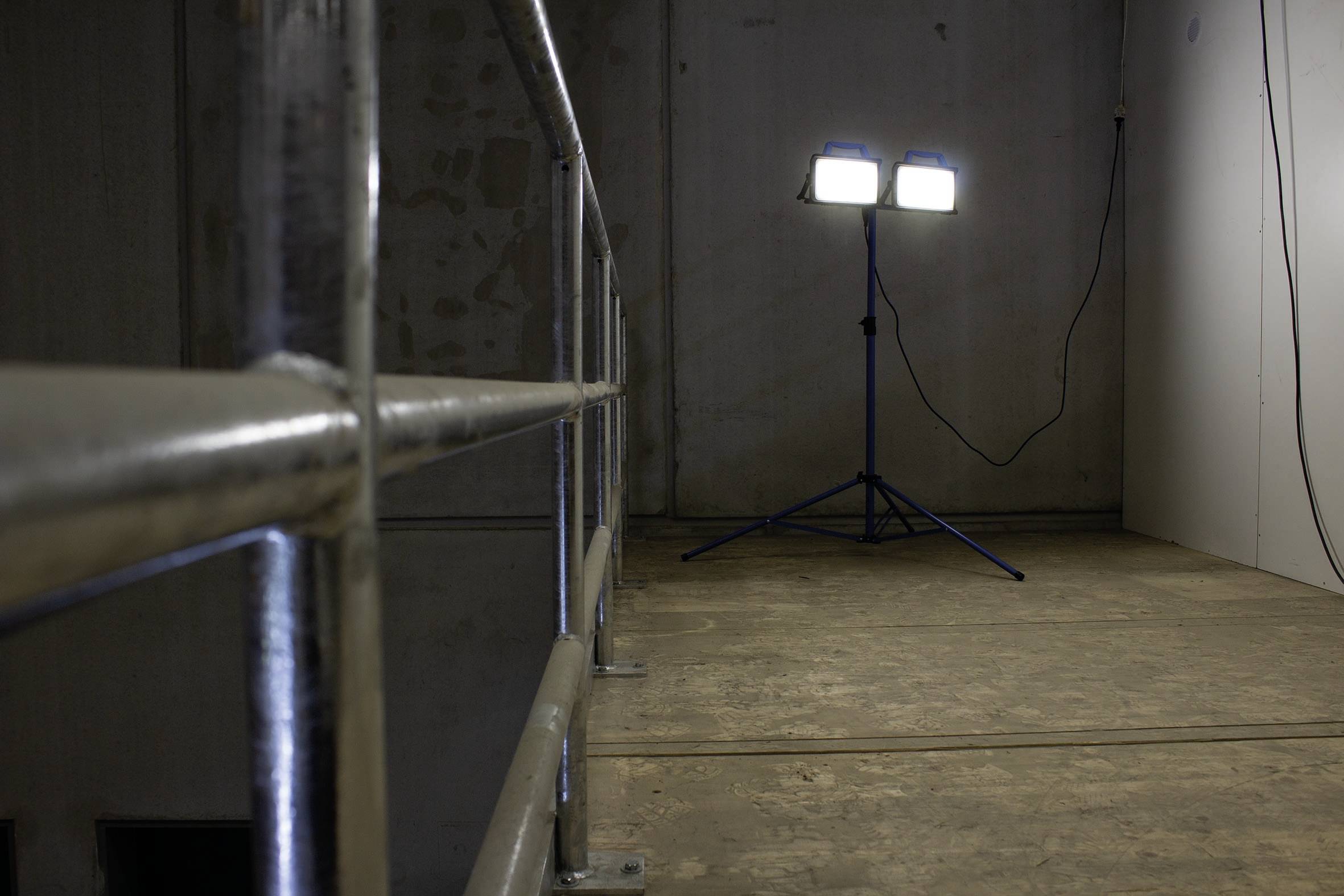 Lešenie s bezpečnostným zábradlím v slabo osvetlenej miestnosti. V rohu stoja dva jasné stavebné reflektory.