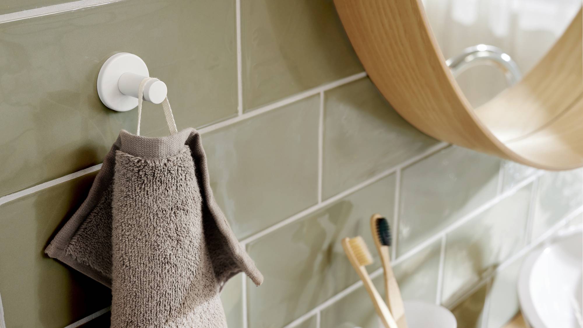 Hnedý uterák visí na bielom háčiku na zelených obkladačkách. V pozadí je zrkadlo a dve drevené zubné kefky v pohári na zubnú hygienu.