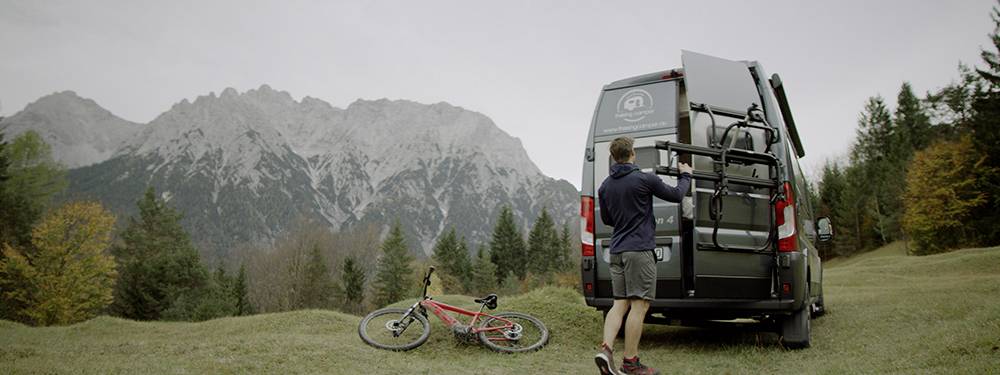 Osoba stojí vedľa dodávky na lúke v horách a ukladá bicykel. V pozadí sú stromy a horský masív.