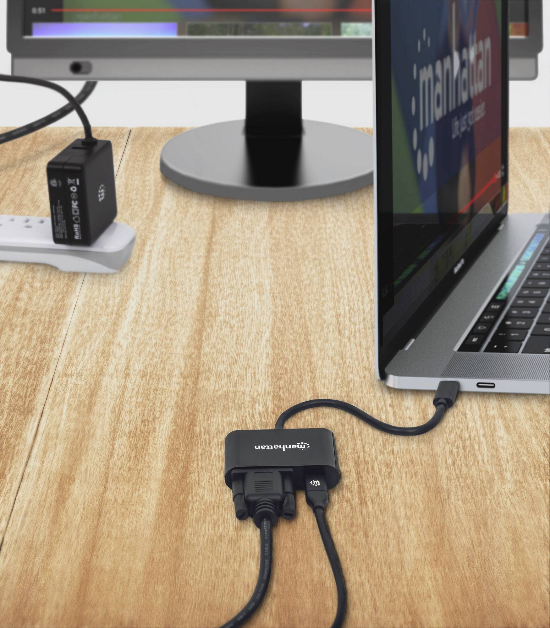Laptop je pripojený cez USB hub na drevenom stole k viacerým zariadeniam. V pozadí stojí monitor.