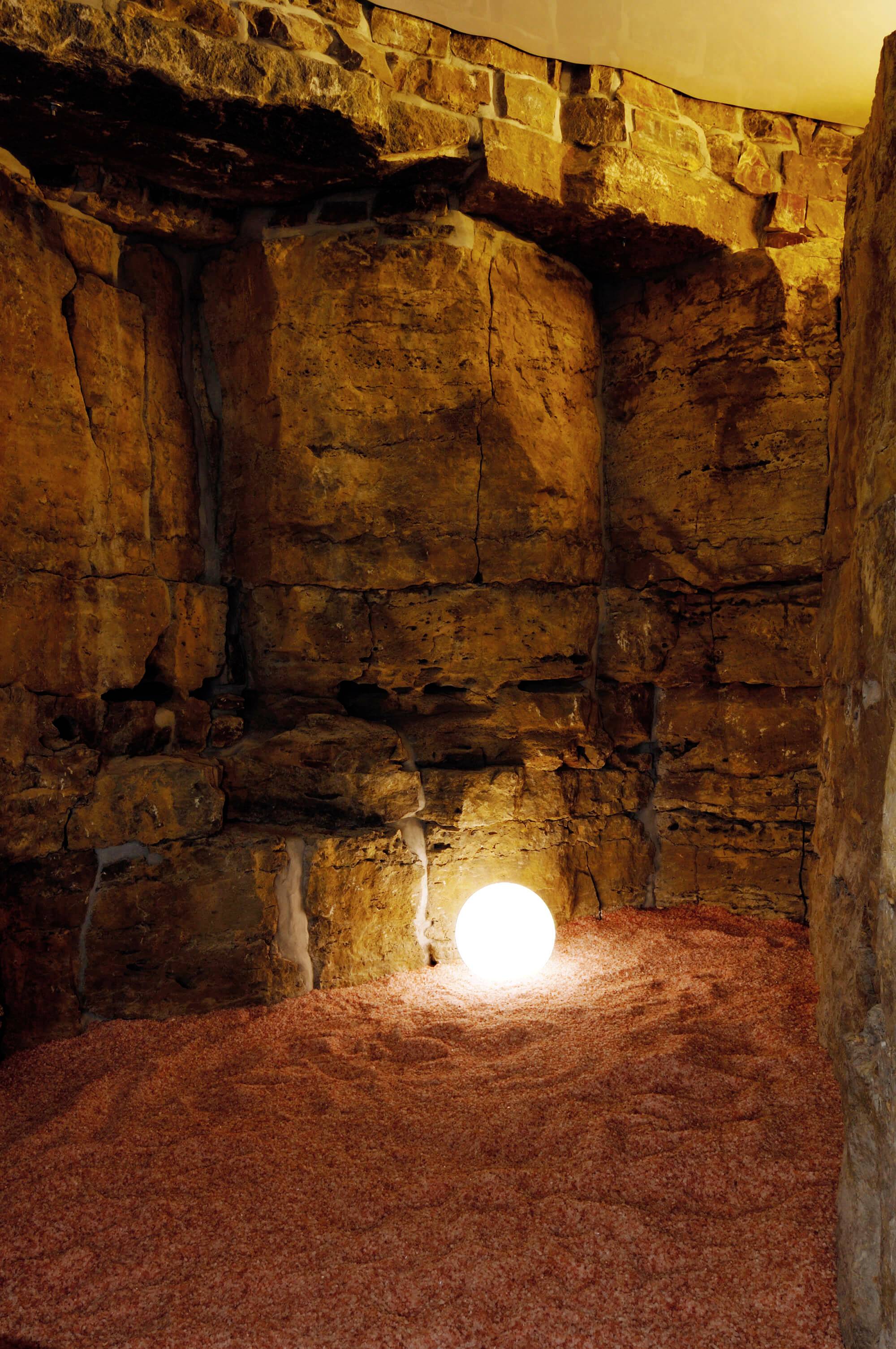 Hrubý, kamenistý priestor s žiarivou guľou na podlahe, ktorá priestor zalievala teplým, žltkastým svetlom.