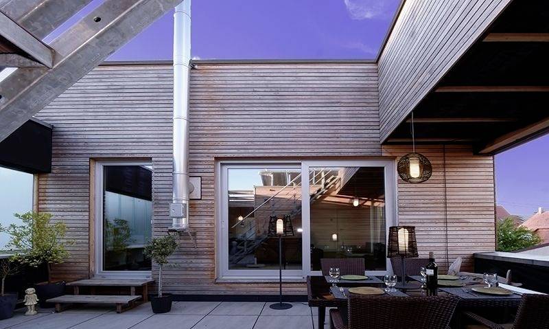 Terasa na modernej plochej streche s drevenými stenami, jedálenským stolom a stoličkami. Obloha je jasná a zafarbená do fialova.