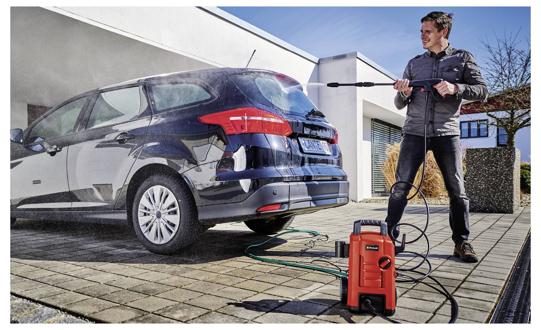 Muž čistí tmavý modrý kombík na príjazdovej ceste pomocou červeného elektrického vysokotlakového čističa.