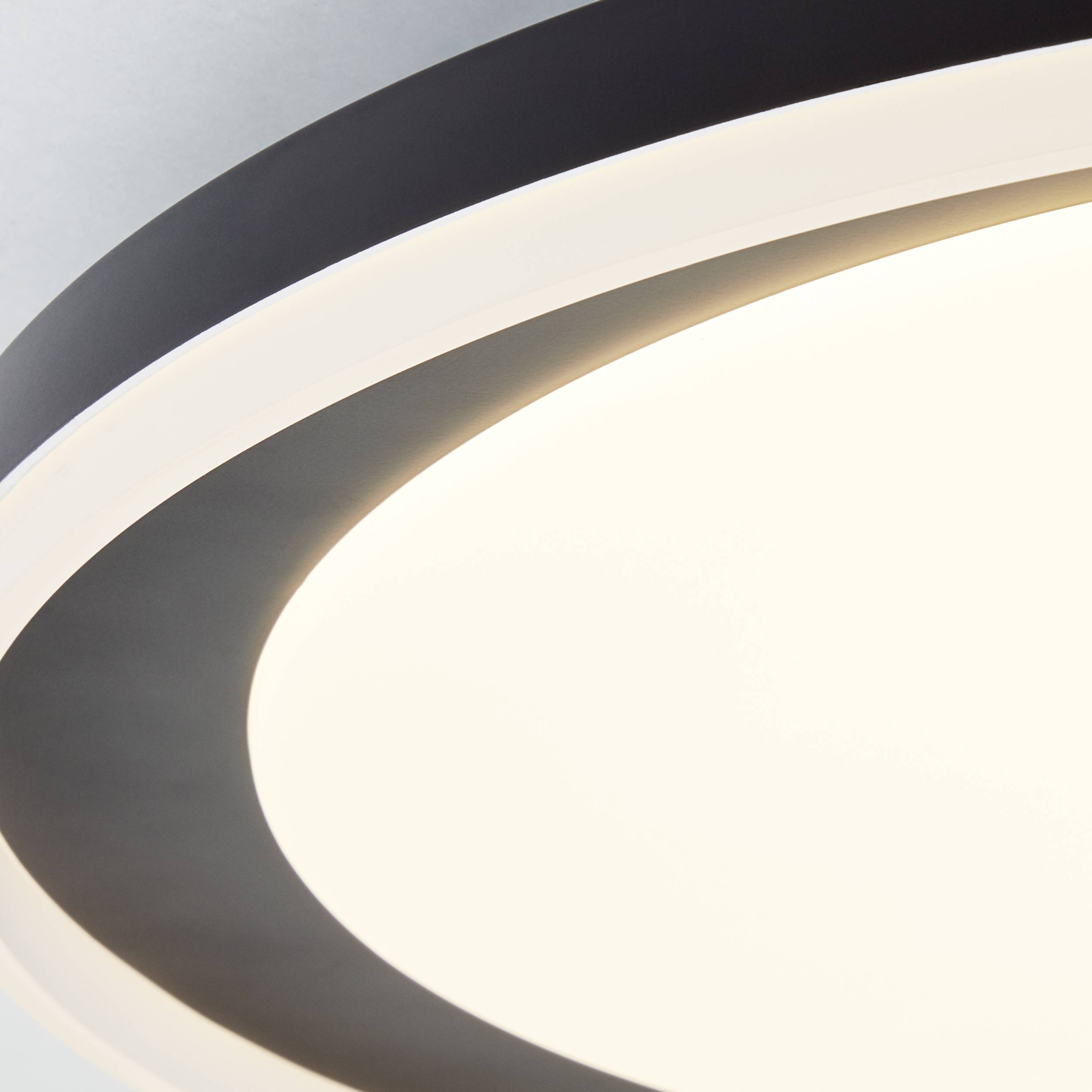 Guľaté, moderné stropné svietidlo svieti bielym svetlom, obklopené čiernym lemom, na šedom pozadí.