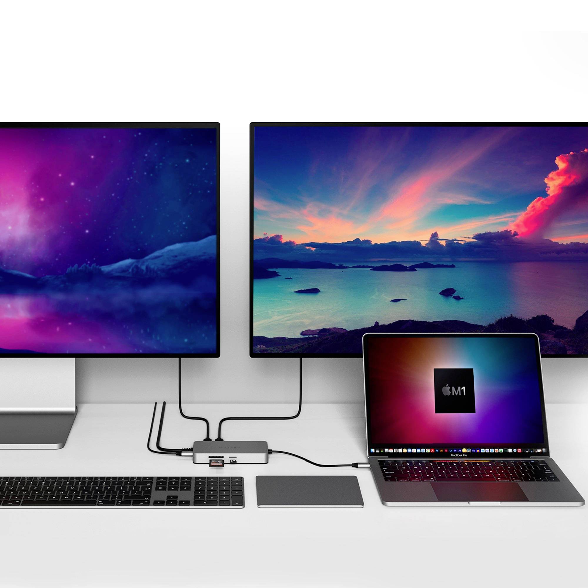 Laptop s čipom M1 je pripojený k dvom veľkým monitorom, ktoré zobrazujú farebné krajinárske obrázky. Pred nimi stojí klávesnica a myš.