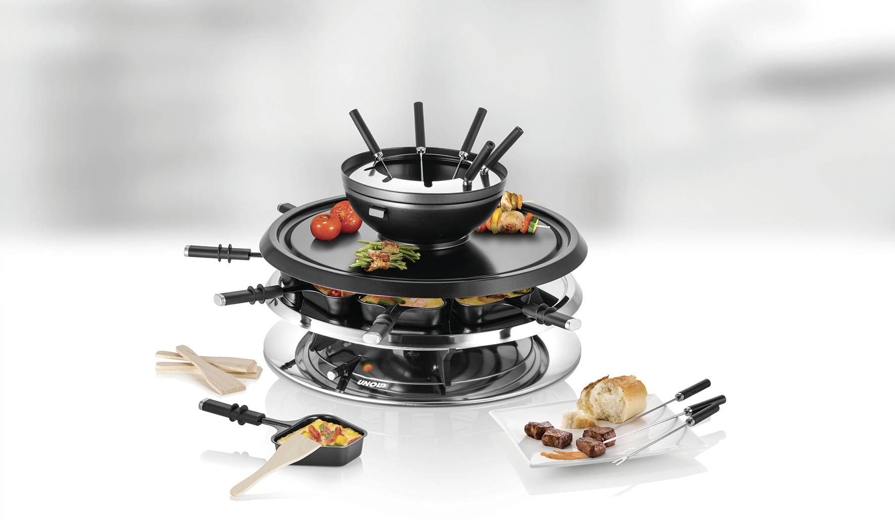 Nehrdzavejúci raclette gril s fondue hrncom, malými panvičkami, zeleninou, chlebom a kúskami mäsa zobrazený na bielom pozadí.