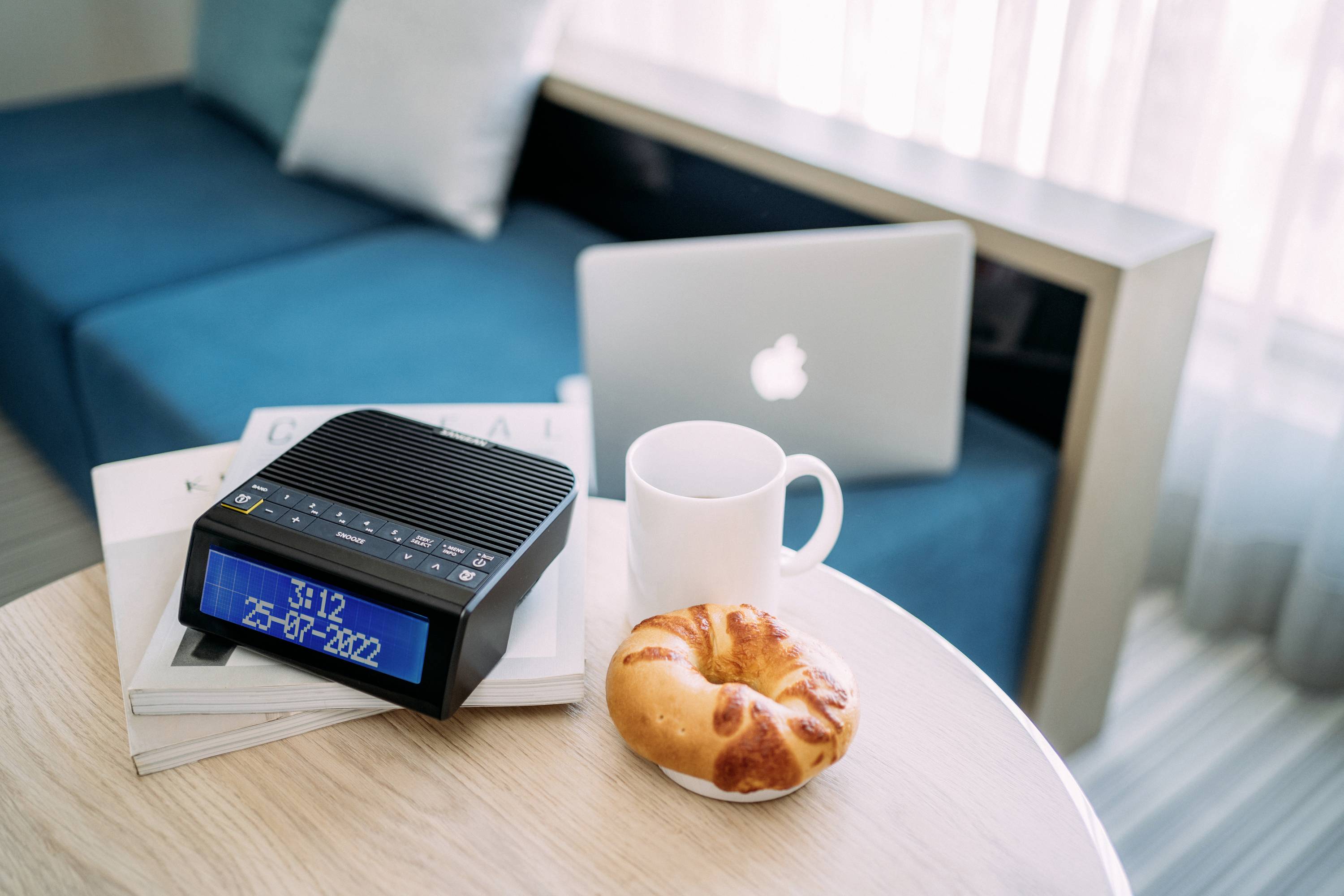 Stôl s kávou, croissantom, digitálnym budíkom a laptopom vedľa gauča. Čas na budíku: 9:12. Útulný pracovný kútik v obývacej izbe.