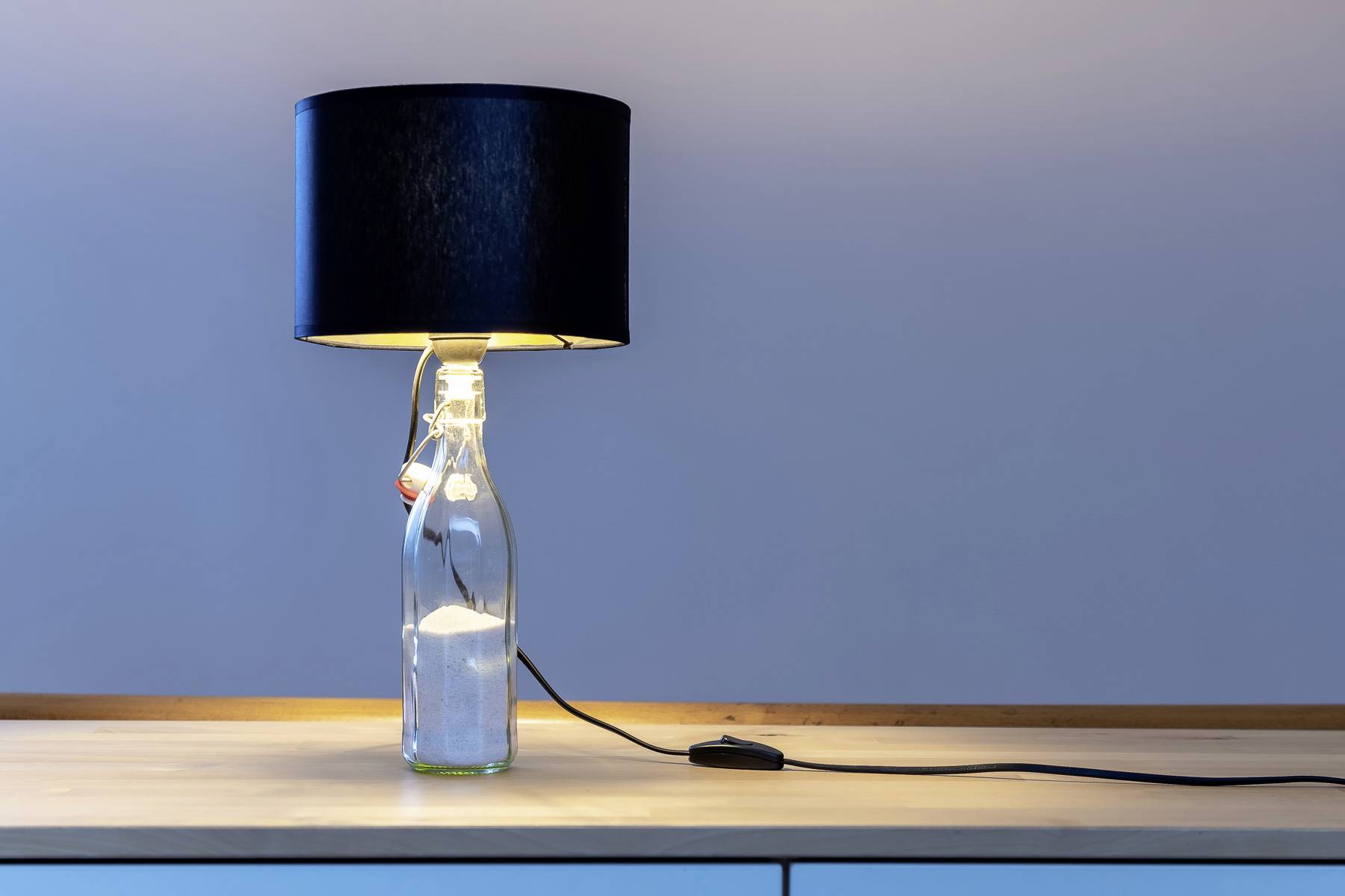 Stolová lampa s čiernym tienidlom, ktorej podstavec tvorí sklenená fľaša, stojí na svetlom drevenom stole.