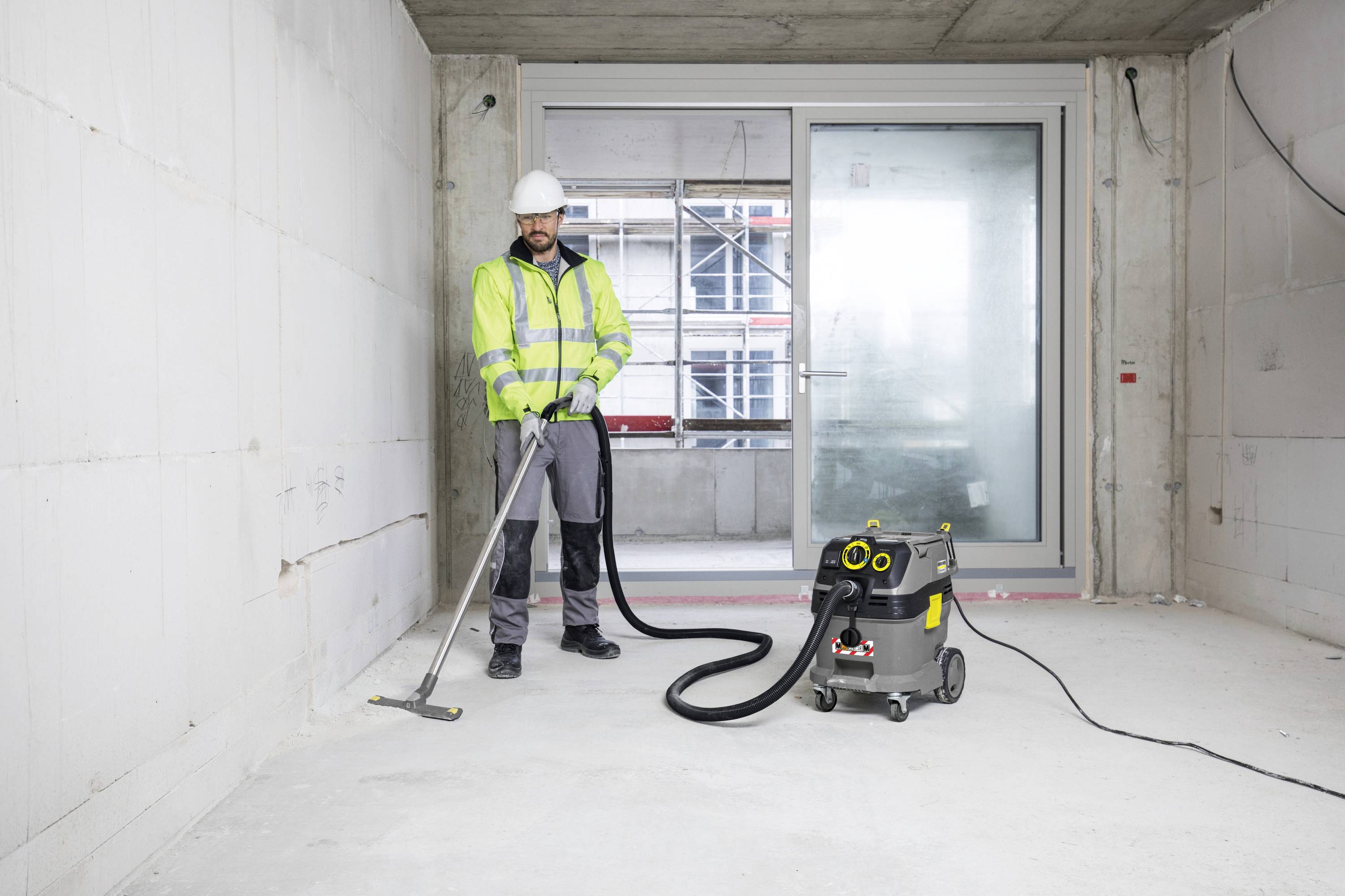 Robotník v ochrannom odeve čistí prázdnu miestnosť priemyselným vysávačom. V pozadí sú veľké okná.