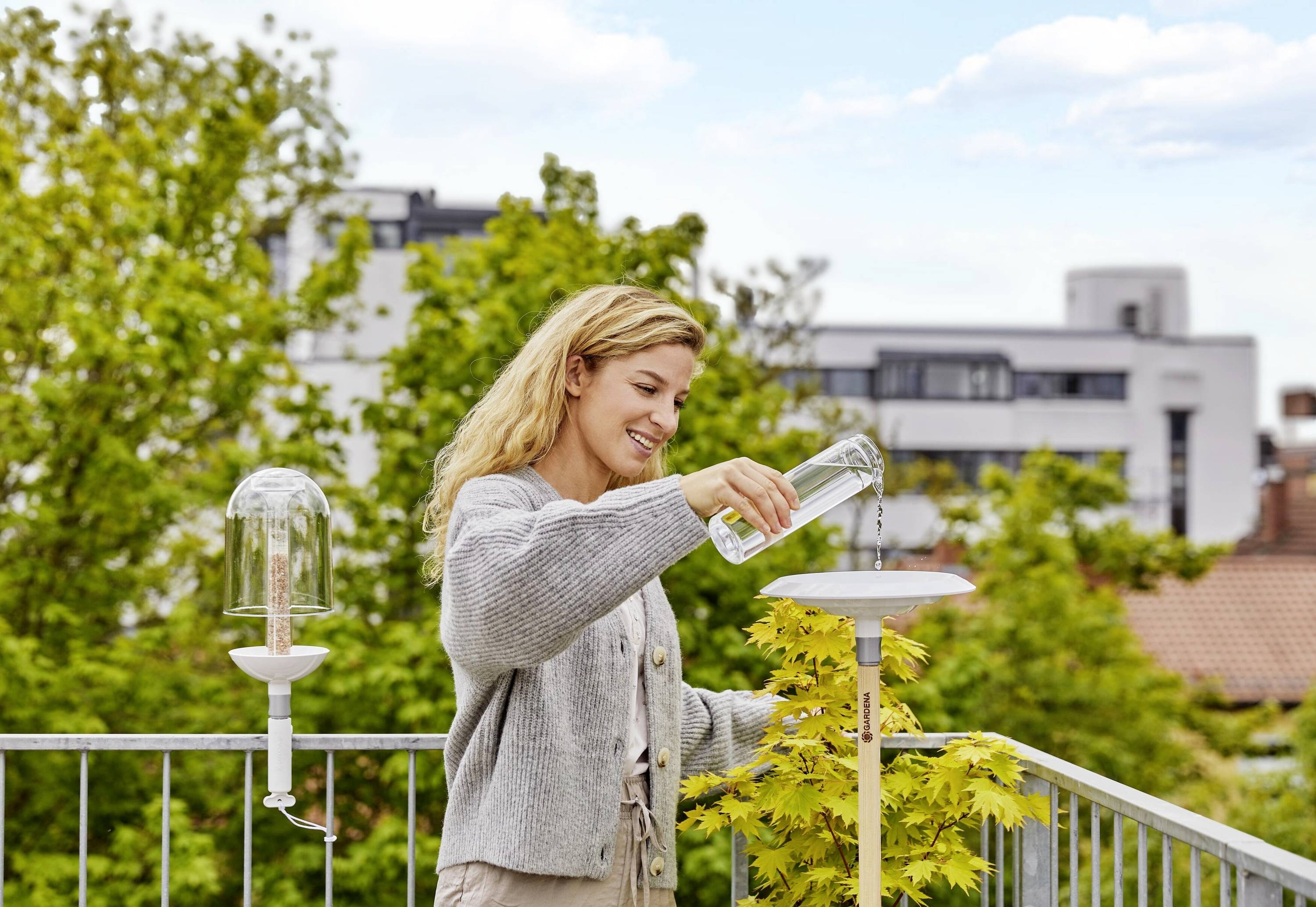 Žena zalievajúca rastlinu na balkóne. V pozadí sú viditeľné stromy a budovy.