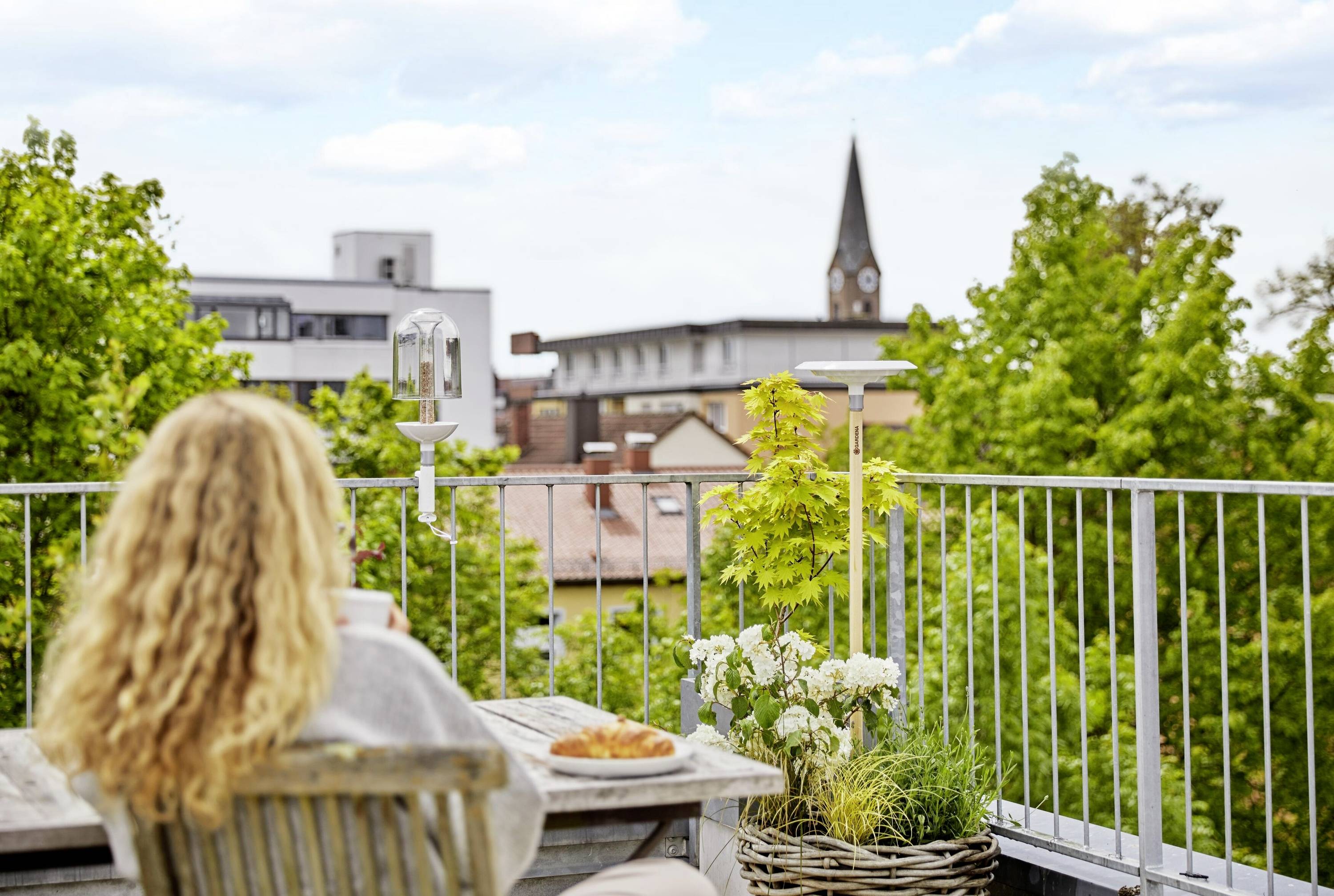 Žena s kučeravými vlasmi sedí na balkóne a díva sa do diaľky. Pred ňou stôl s kávou a pečivom. V okolí zelené stromy, v pozadí kostolná veža.