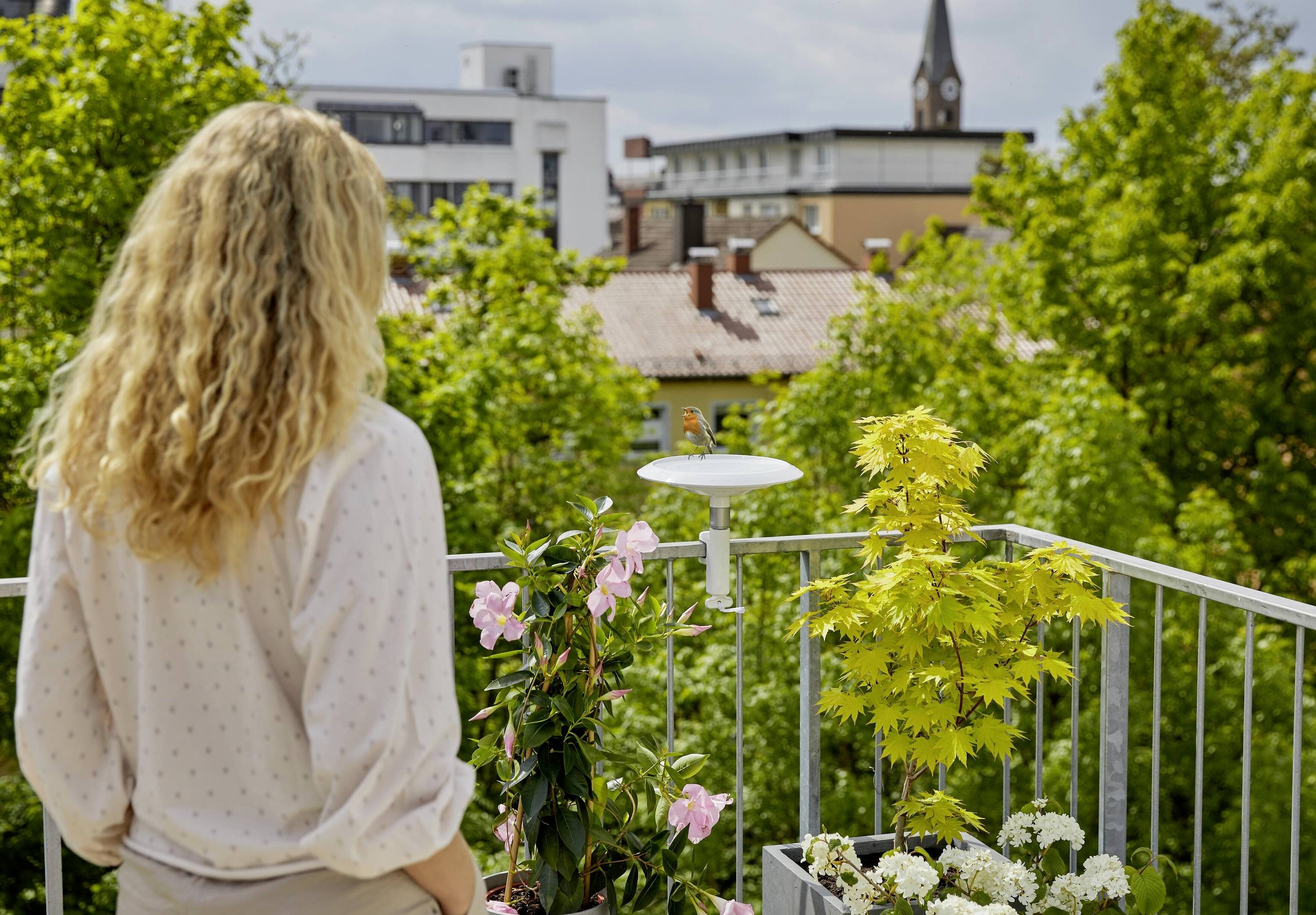 Osoba s dlhými vlasmi stojí na balkóne, obklopená kvitnúcimi rastlinami. V pozadí sú viditeľné stromy a budovy.