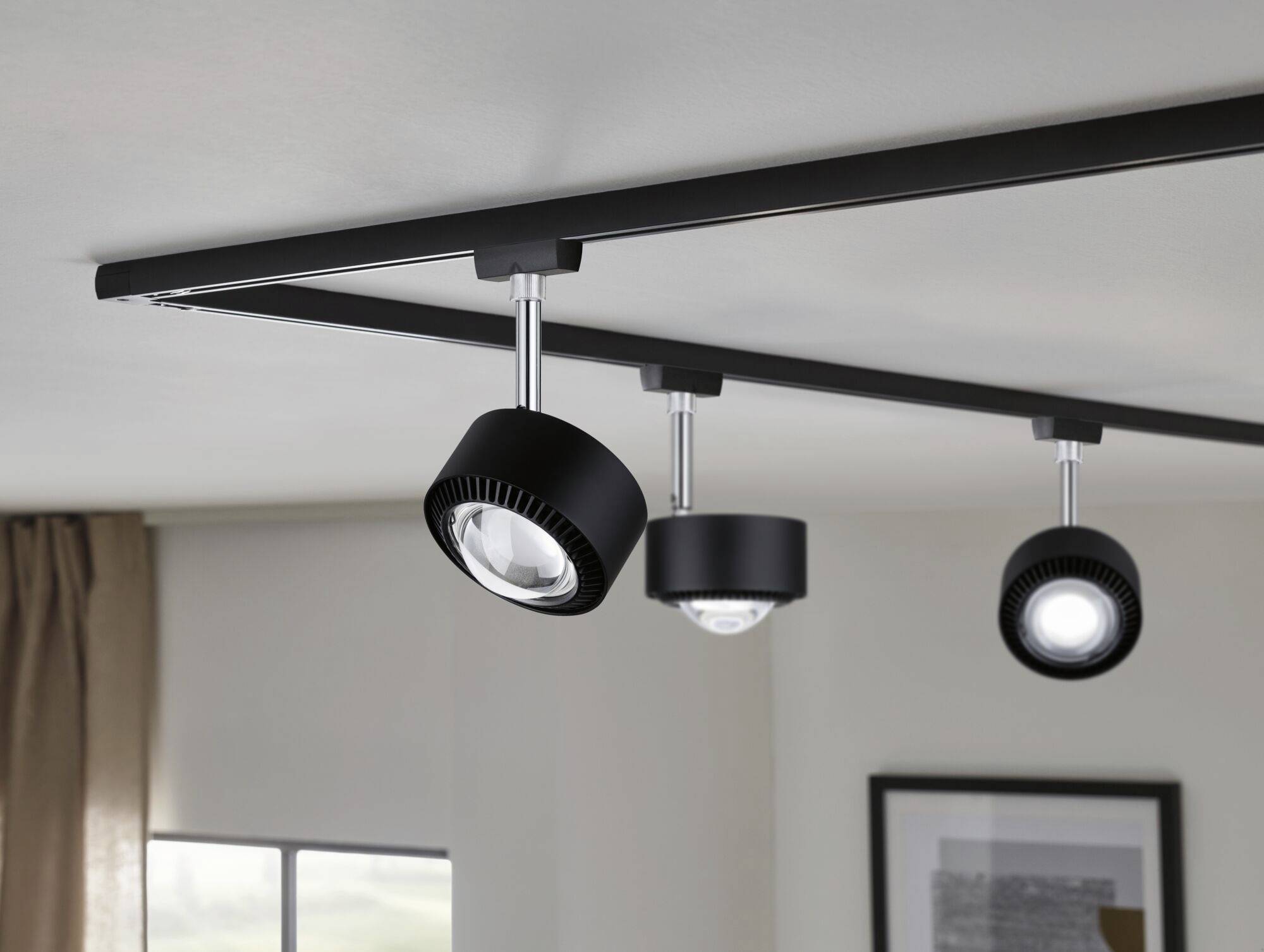 Moderná čierna stropná lampa s tromi nastaviteľnými reflektormi, namontovaná na koľajnici, osvetľuje obytný priestor.