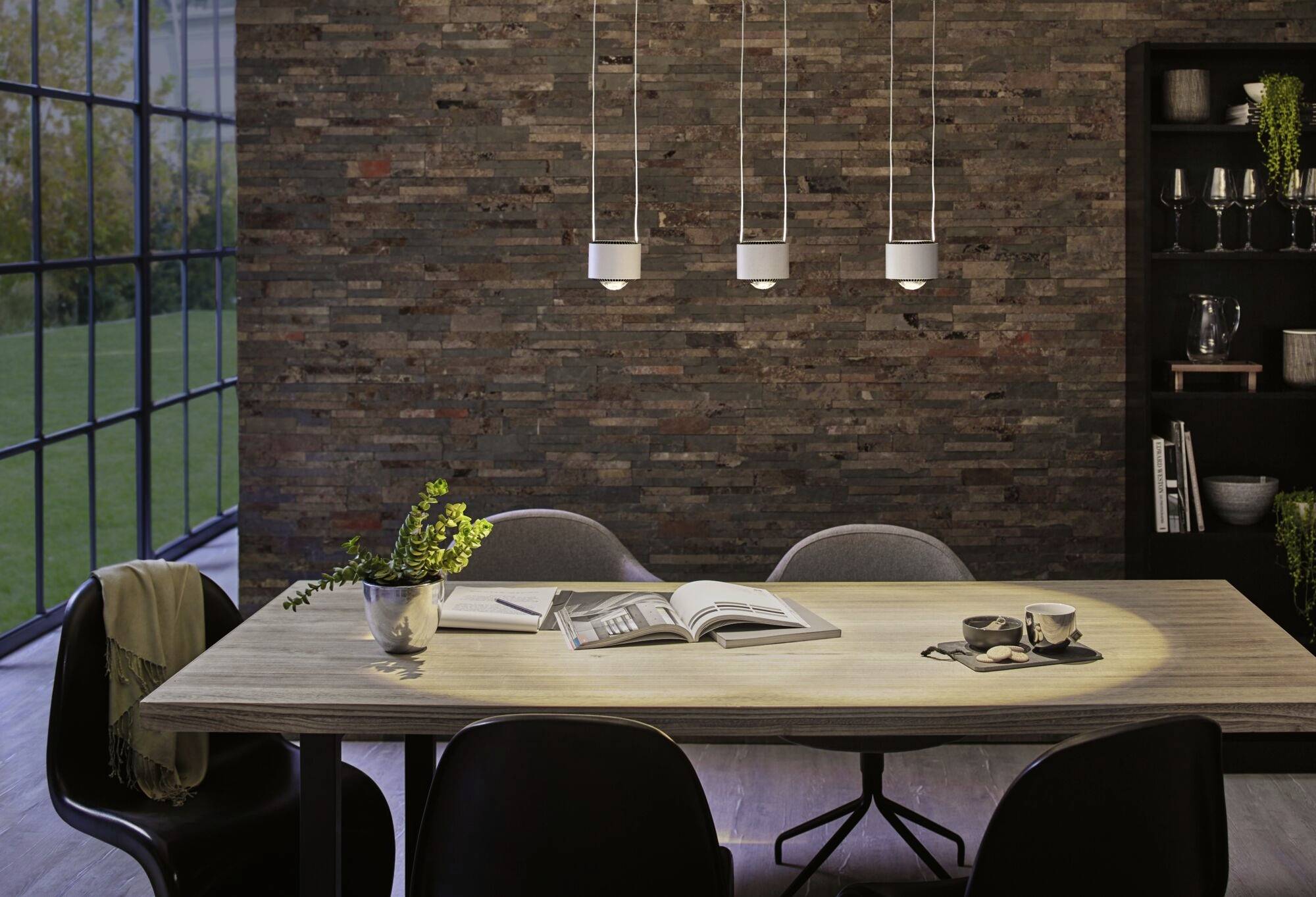 Jedáleň s dreveným laminátovoým stolom, tromi závesnými svetlami, štyrmi stoličkami, otvorenými knihami, dvomi šálkami a rastlinou v kovovom kvetináči.