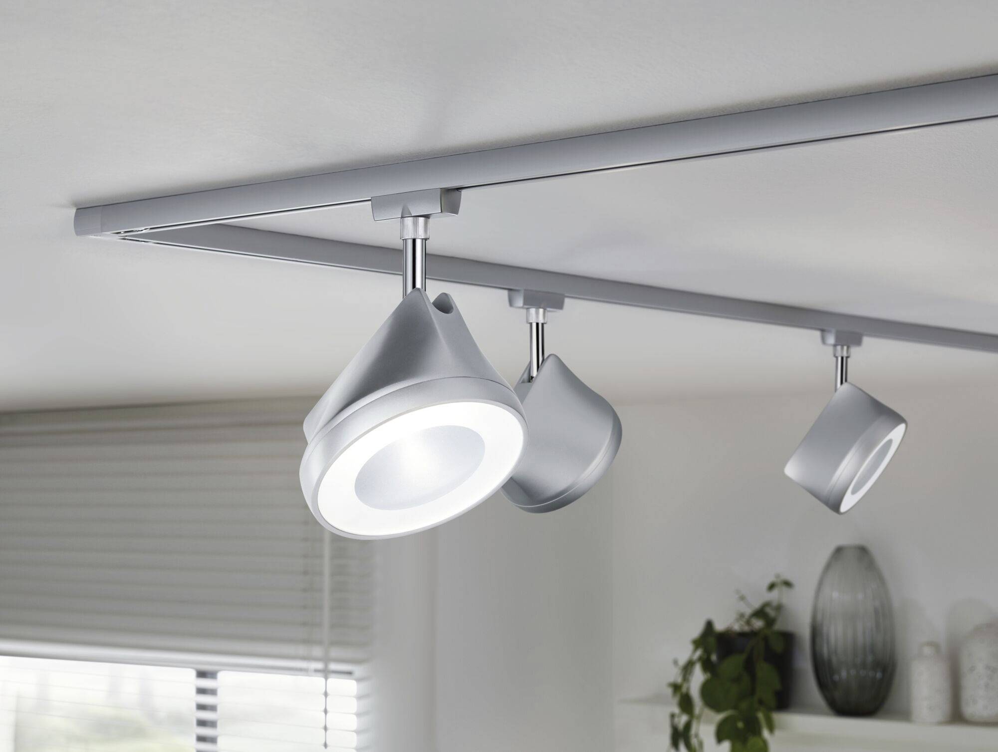 Tri moderné strieborné stropné svietidlá namontované na lište osvetľujú priestor. V pozadí sú viditeľné rastliny a dekorácie.