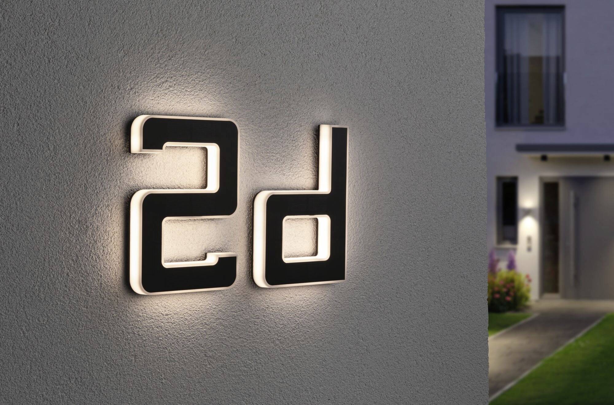 Svietiace domové číslo '2d' na sivej stene domu; v pozadí rozmazane viditeľná záhrada a vstup do domu.