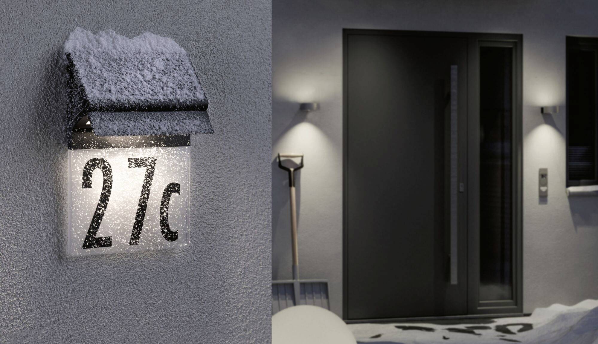 Číslo domu '27c' osvetlené na zasneženom múre. Vpravo moderný vstup so súpravou na odhŕňanie snehu vedľa neho.