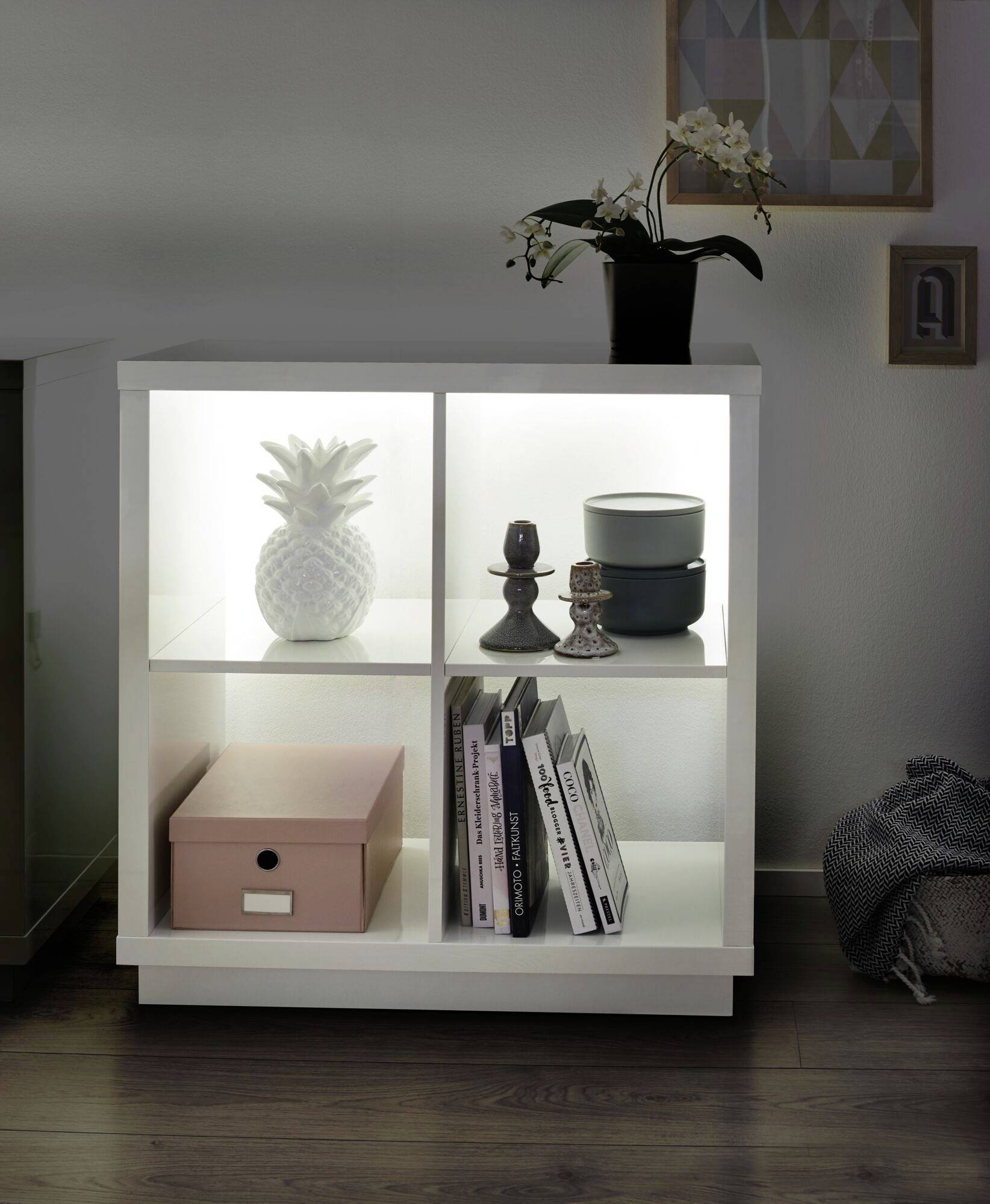 Biela polica so štyrmi osvetlenými priehradkami obsahuje figúrku ananásu, svietniky, škatuľu, knihy a orchideu.