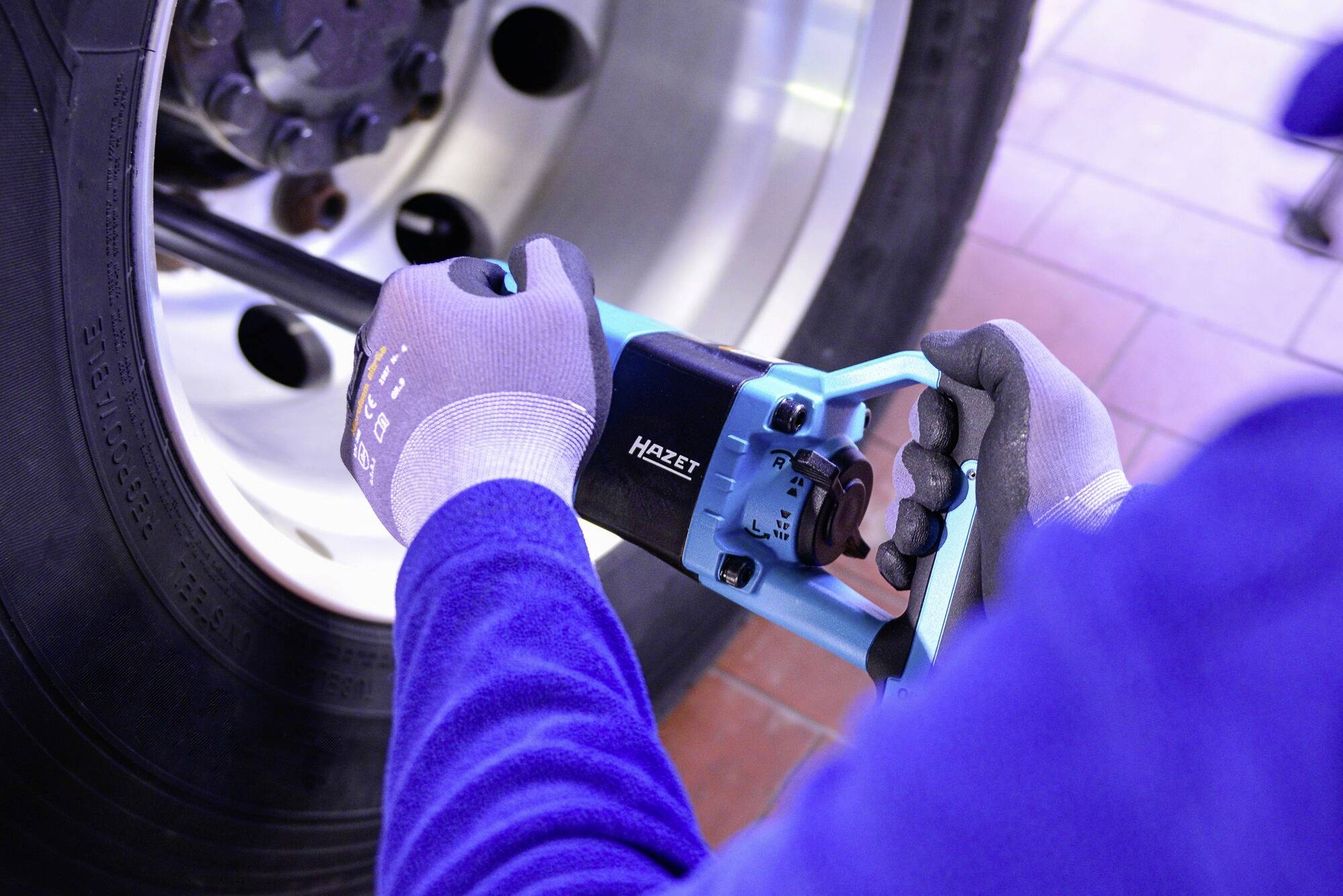 Osoba s modrým náradím uťahuje skrutky na veľkej pneumatike. Má na sebe pracovné rukavice.