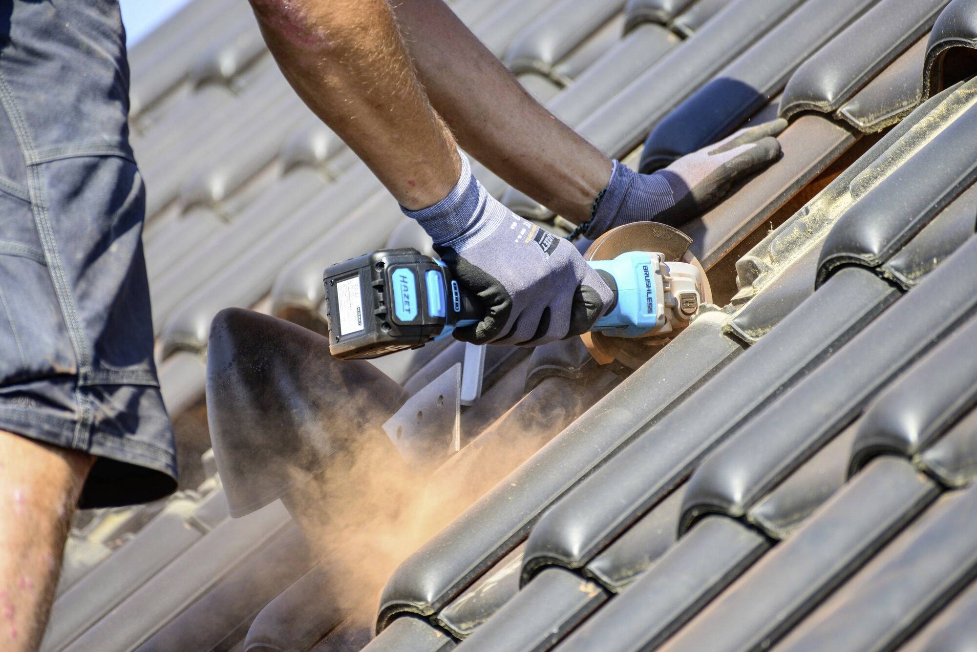 Robotník v krátkych nohaviciach a rukaviciach používa elektrické náradie na rezanie tehál na streche. Prach sa vznáša, slnečné počasie.