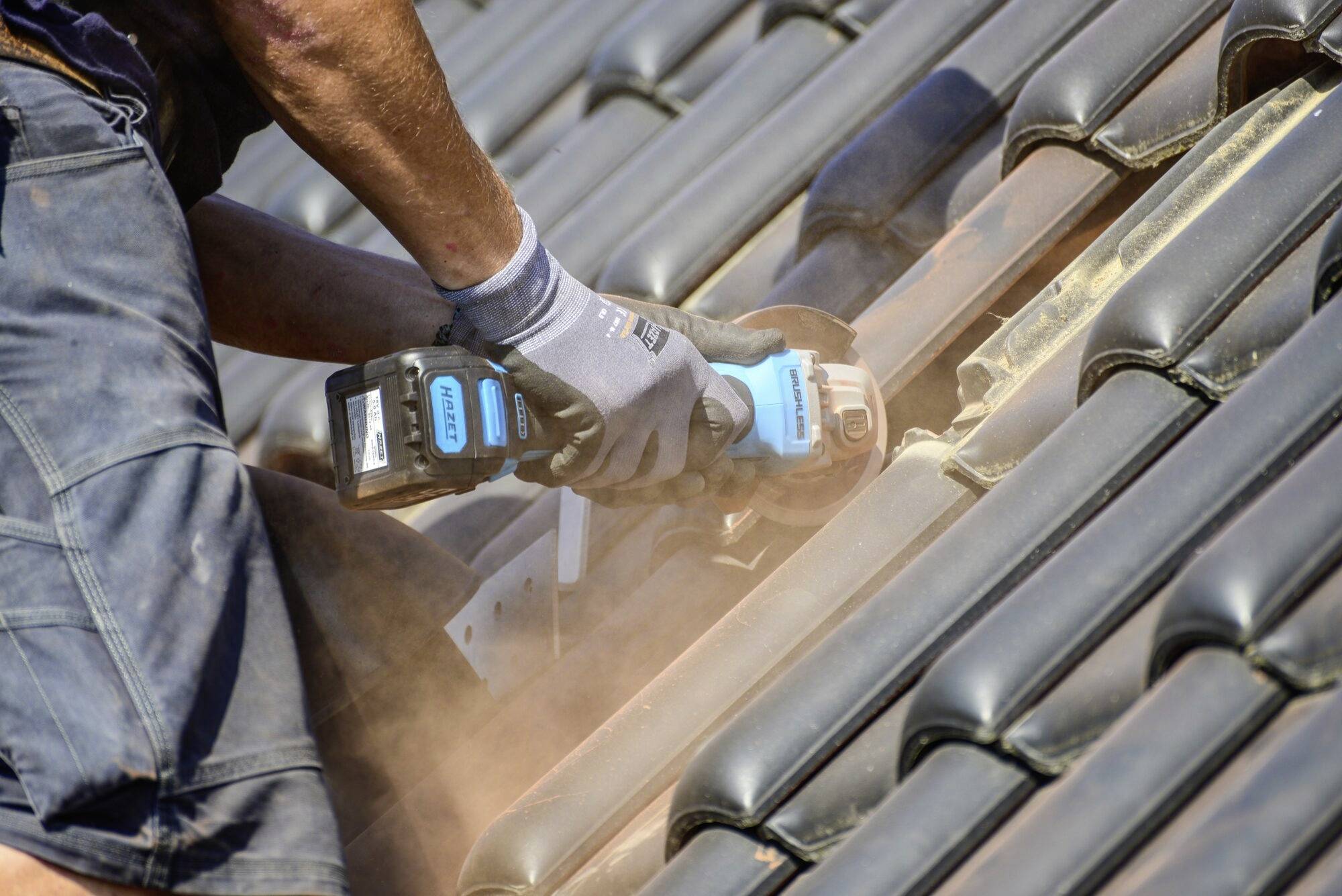 Osoba pracuje s elektrickou pílou na streche pokrytej čiernymi škridlami. Z rezaných škridiel sa dvíha prach.