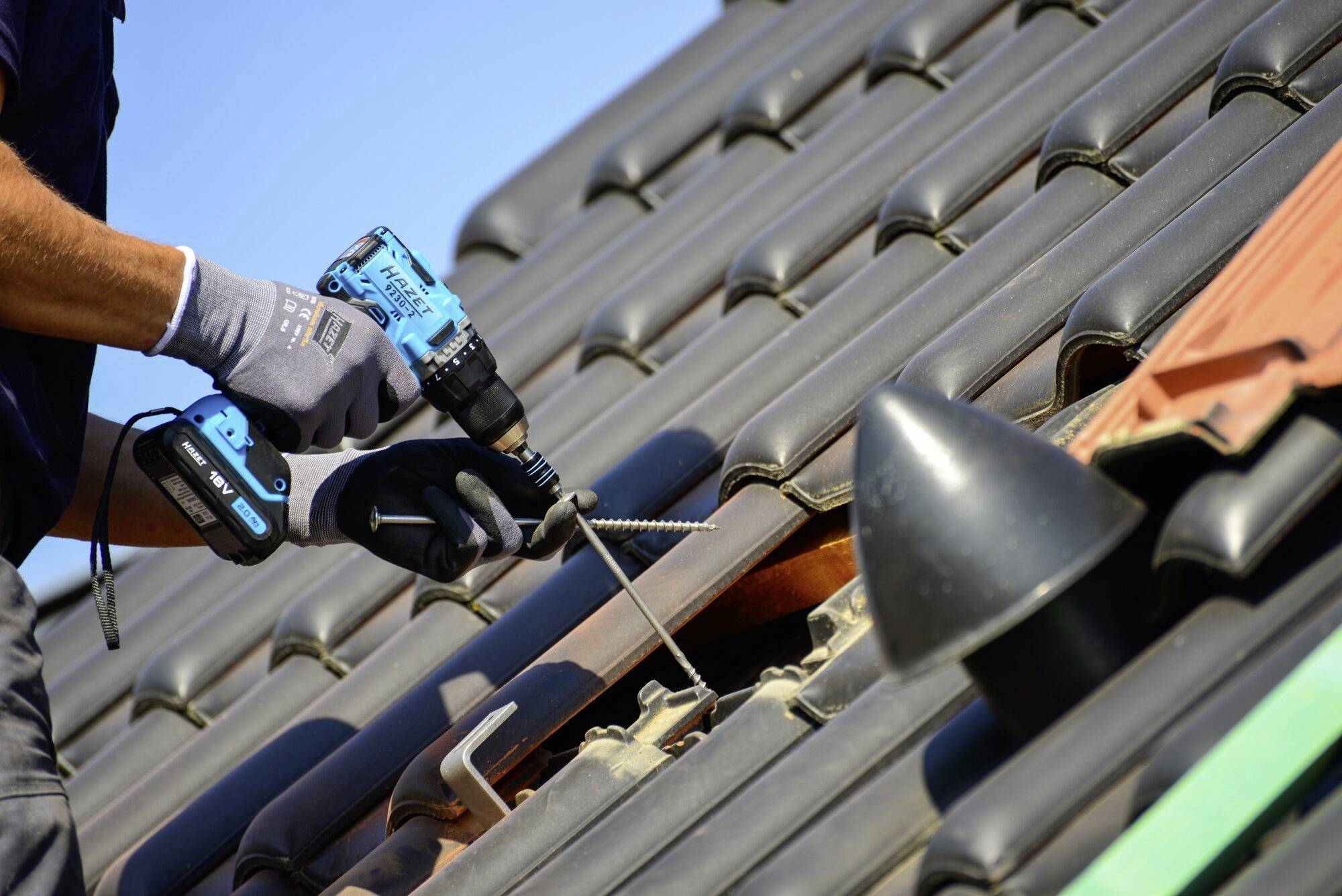 Osoba na streche montuje solárnu elektráreň pomocou akumulátorového skrutkovača. Má na sebe pracovné rukavice.
