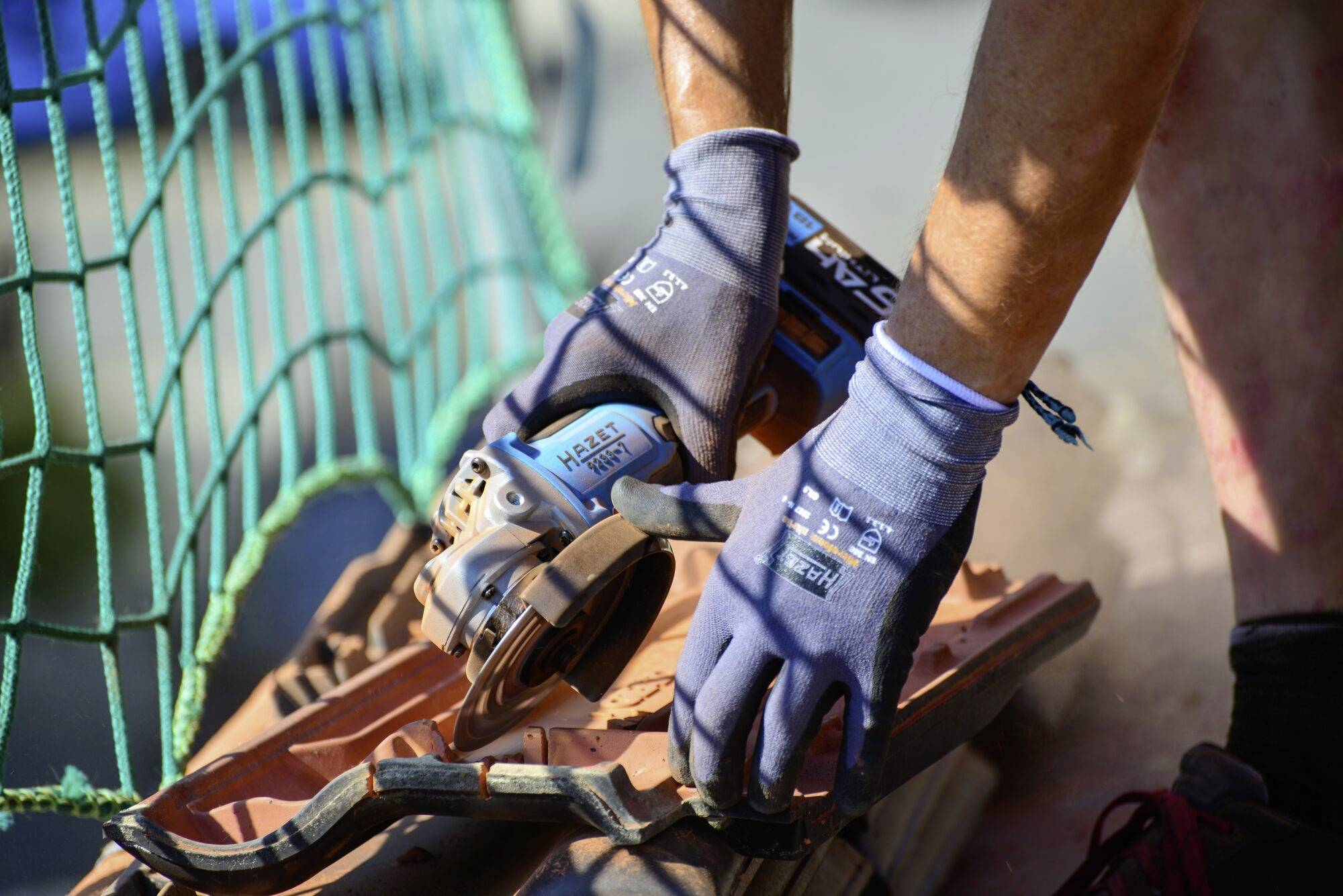 Osoba v rukaviciach striháva škridlu elektrickým nástrojom. V pozadí je vidieť zelené ochranné siete.