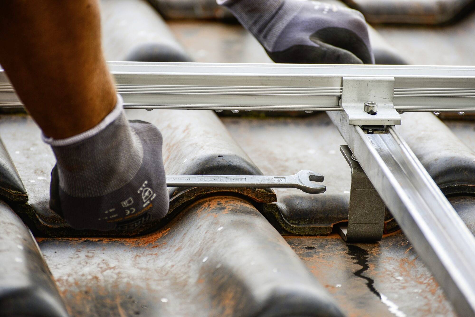 Osoba upevňuje kovovú podporu na tehlové streche pomocou kľúča, pričom má na rukách pracovné rukavice.