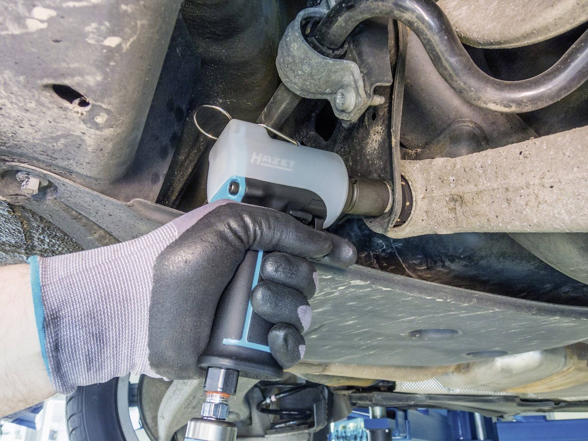 Osoba používa elektrický nástroj na uvoľnenie skrutky na spodnej časti vozidla. Osoba má na rukách rukavice.