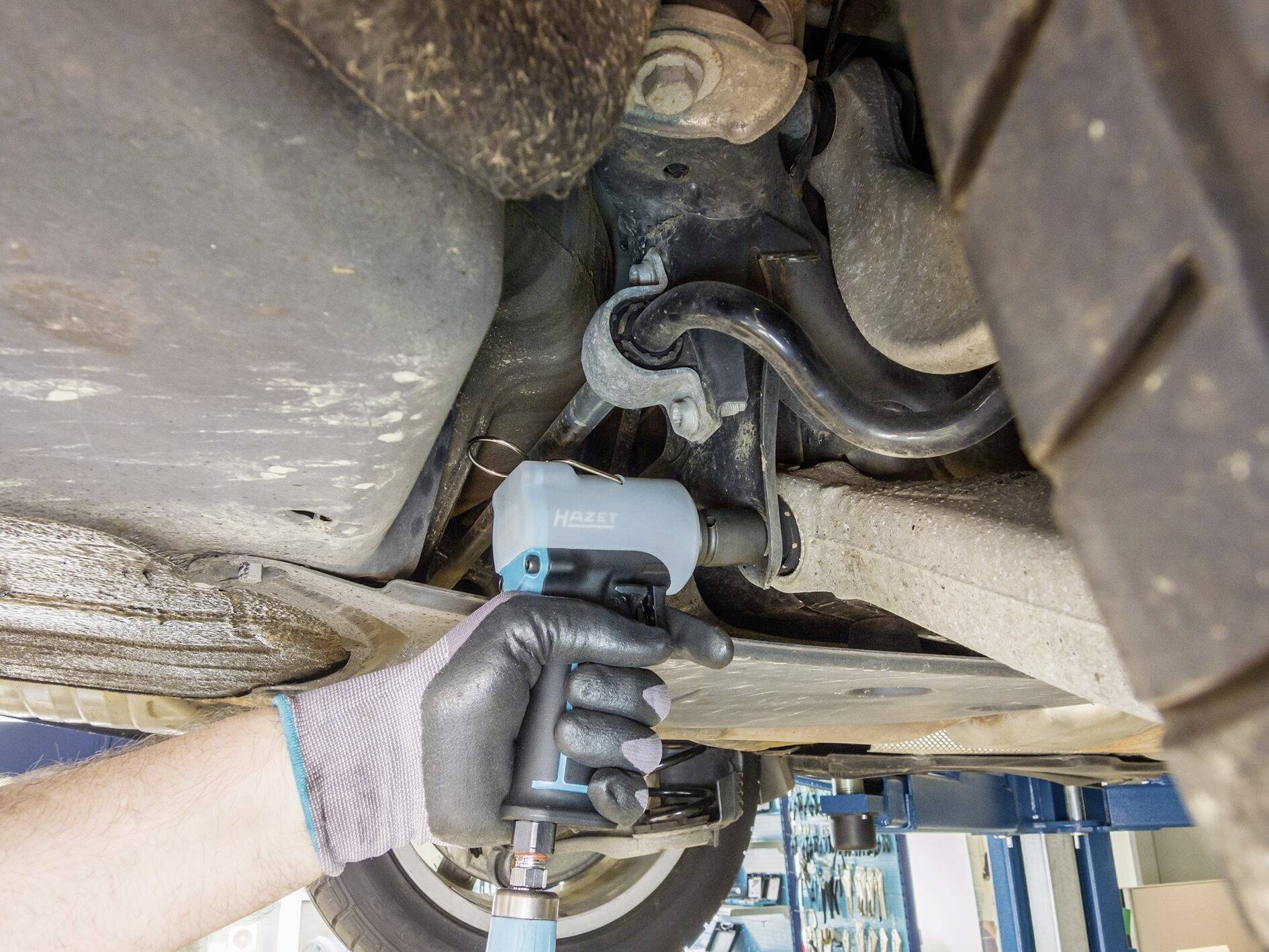 Mechanik v rukaviciach používa pneumatický nástroj na uvoľnenie skrutky na spodnej časti automobilu v autoservise.