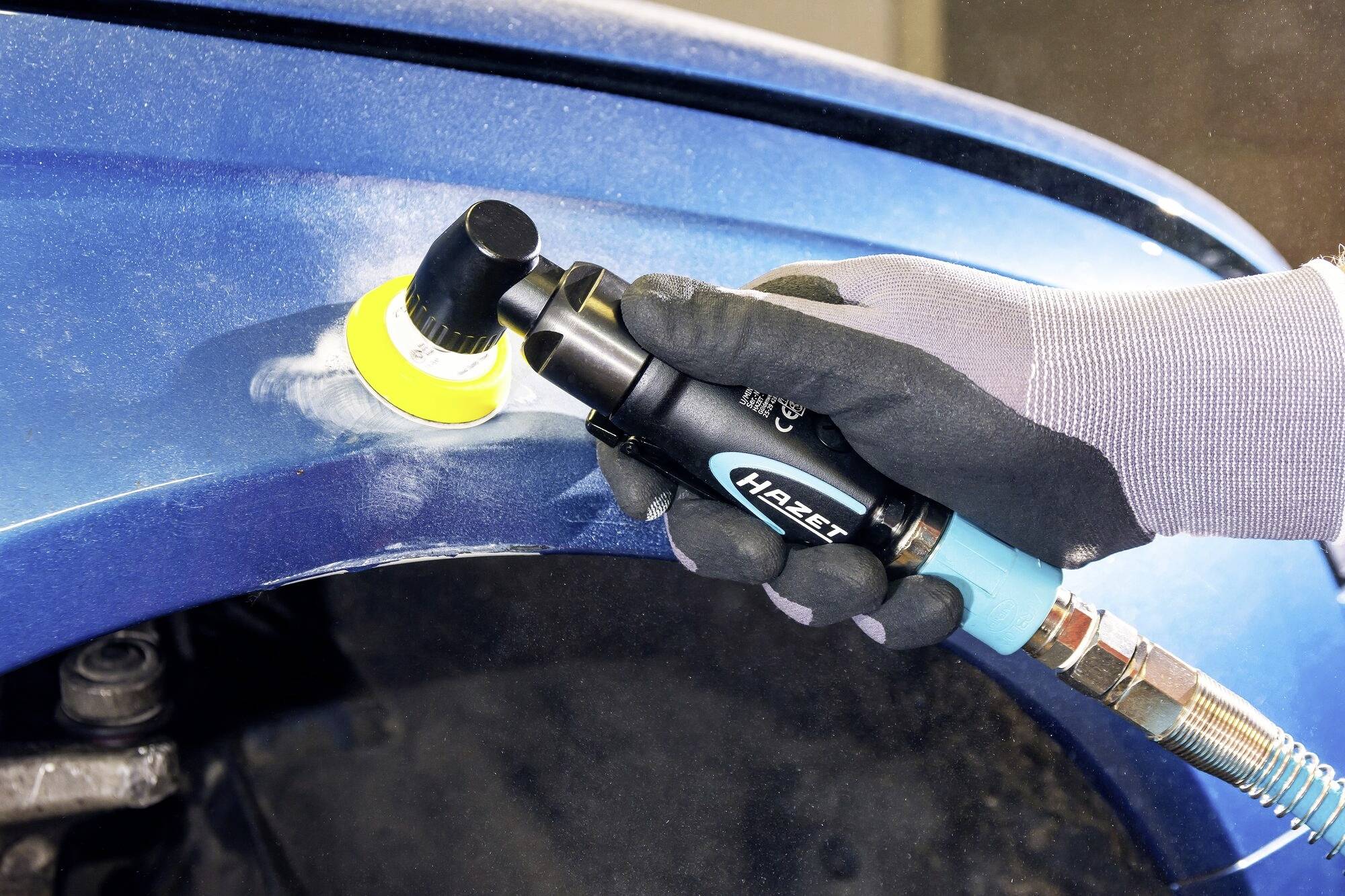 Osoba brúsi modrú karosériu automobilu ručnou brúskou, aby odstránila poškodenia. Má na rukách ochranné rukavice pre bezpečnosť.