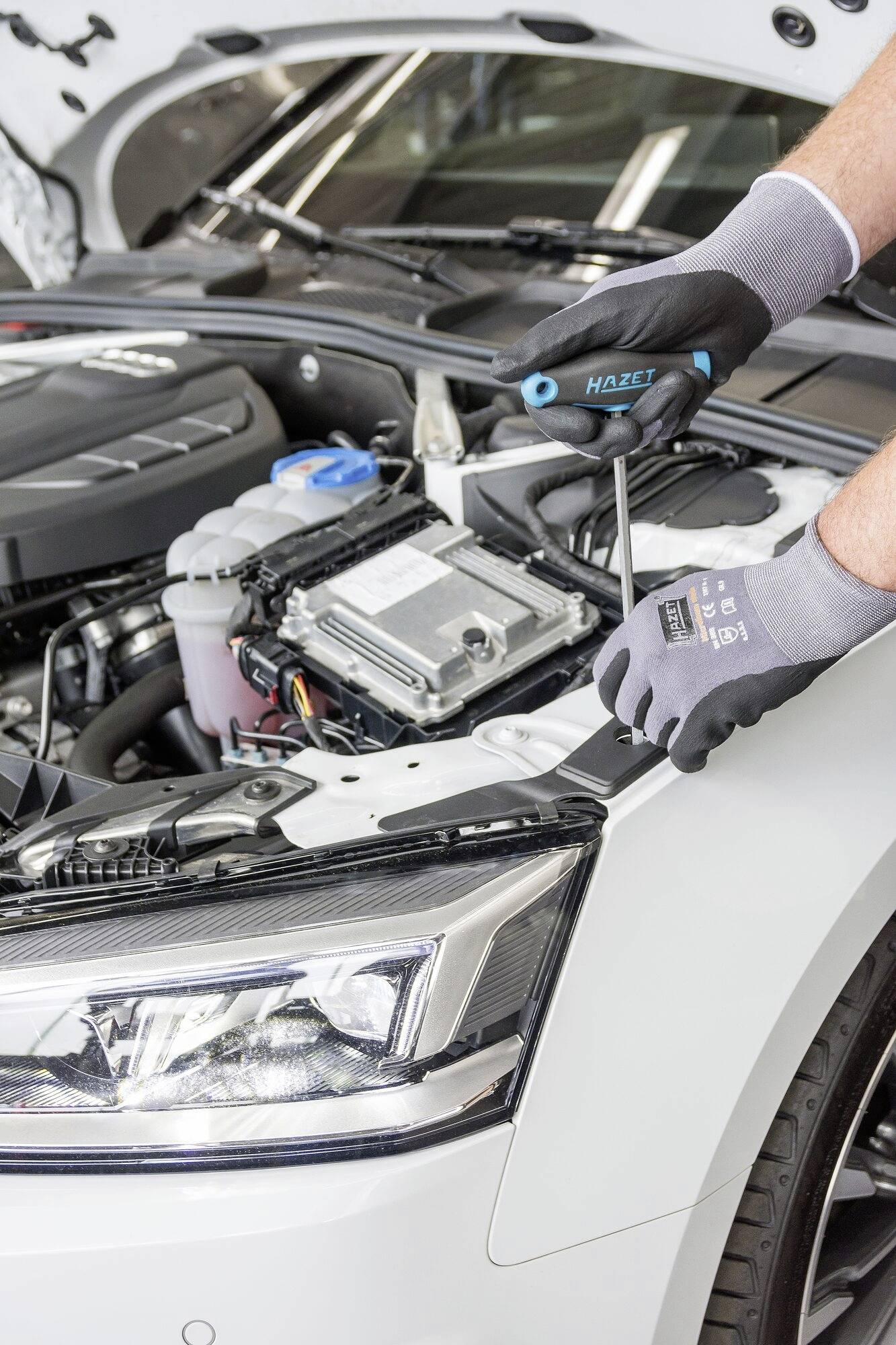 Mechanik pracuje so skrutkovačom v motorovom priestore auta, ktoré stojí v dielni s otvorenou kapotou.