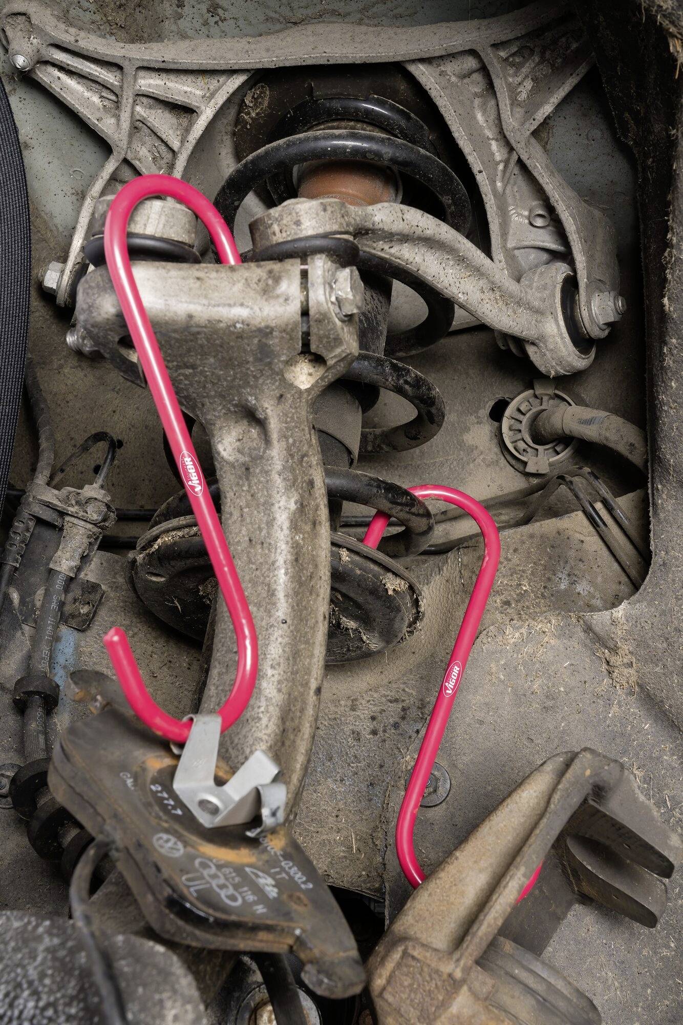 Detailný záber na automobilovú súčiastku s červenými brzdovými strmeňmi a brzdovým kotúčom. Na súčastiach sú viditeľné známky opotrebenia a znečistenia.